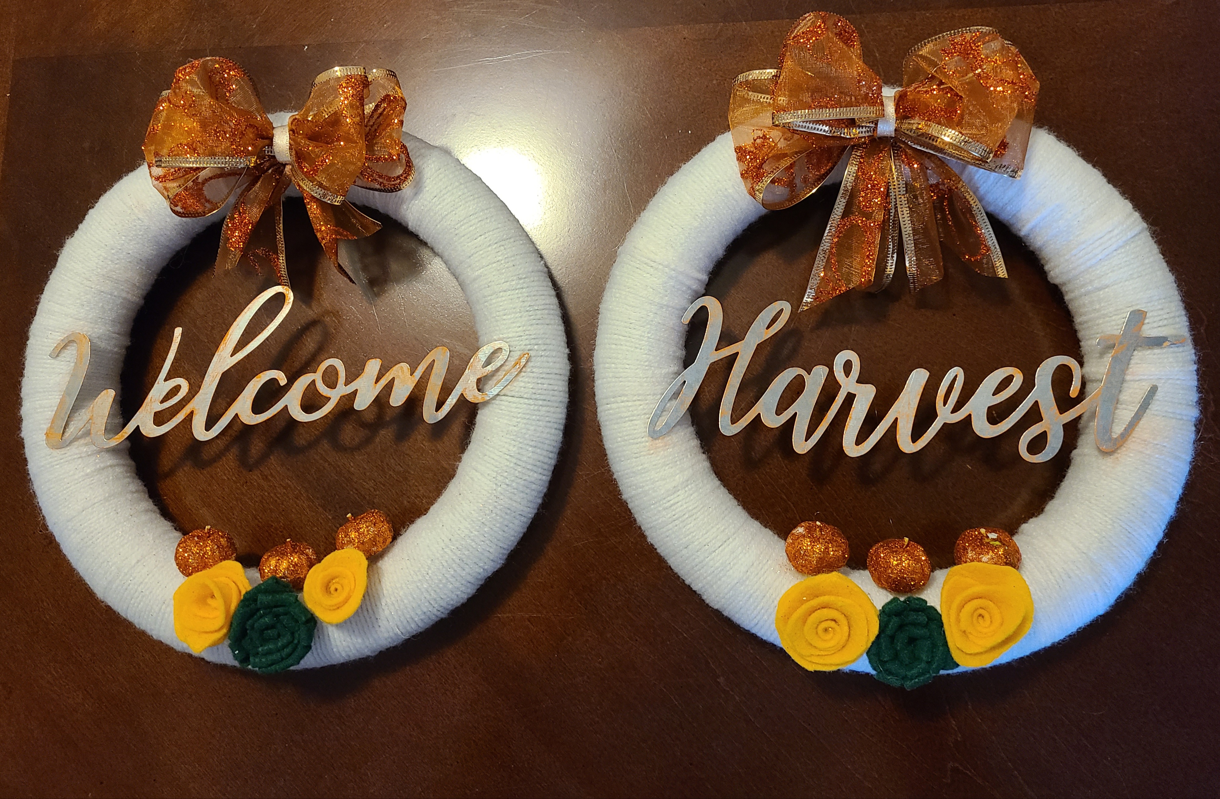 Two completed easy DIY fall wreaths next to each other- styrofoam forms covered with white yarn, orange pumpkin glittered ribbon made into bows placed at the top of each one, metal words in the middle "Welcome" on one "Harvest" on the other. Felt flowers are at the bottom with tiny orange glittered pumpkins glued above them.