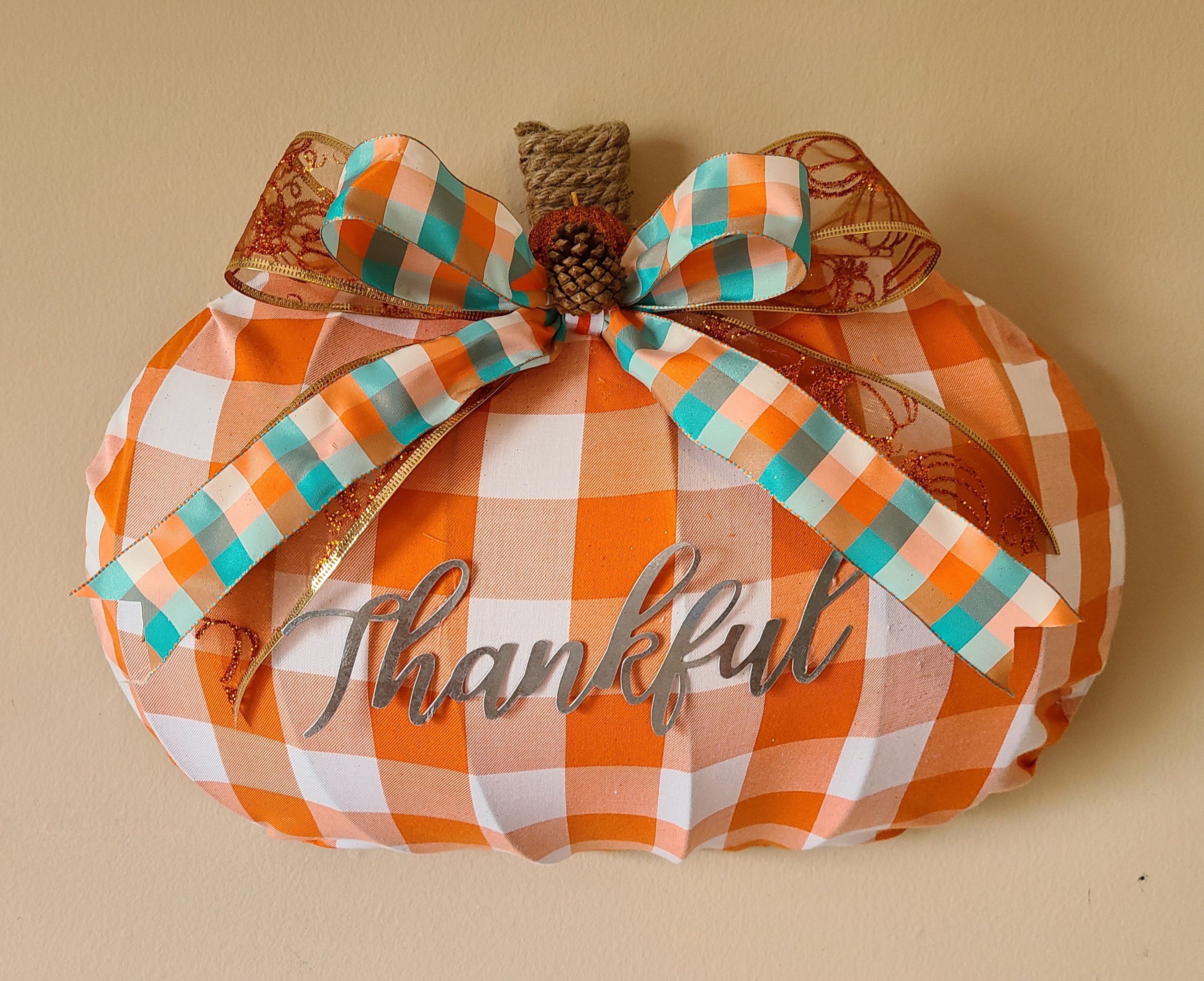 Dollar Tree pumpkin wreath form covered with orange and white buffalo check fabric, galvanized metal "Thankful" in the center, nautical rope wrapped around the top to create a stem, and a teal, orange, & white bow on top of orange and gold glitter ribbon bow.