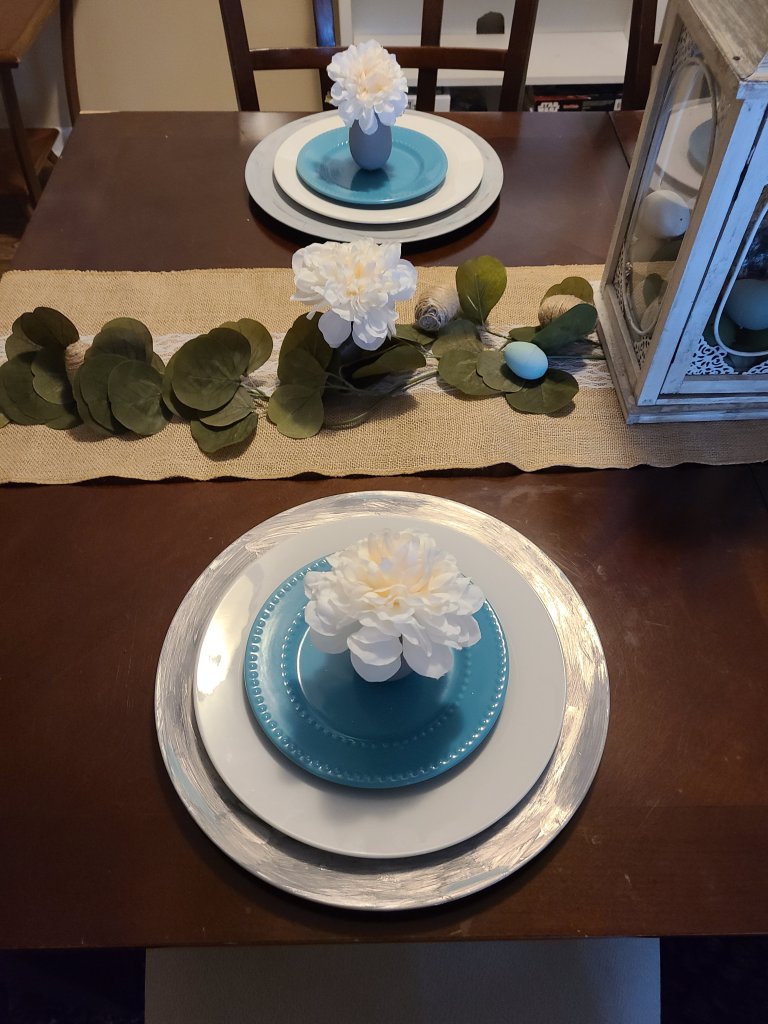 Easter tablescape with burlap and lace table runner and two place settings with gray charger, white plate, and robin's egg blue dessert plate. On top of the plate is an egg vase with white flower in it.
