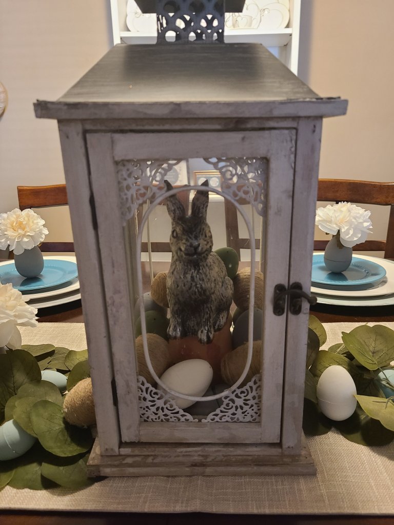 Lantern filled with chalk painted eggs, a terracota pot, & a silver rabbit on top of it on an Easter tablescape.