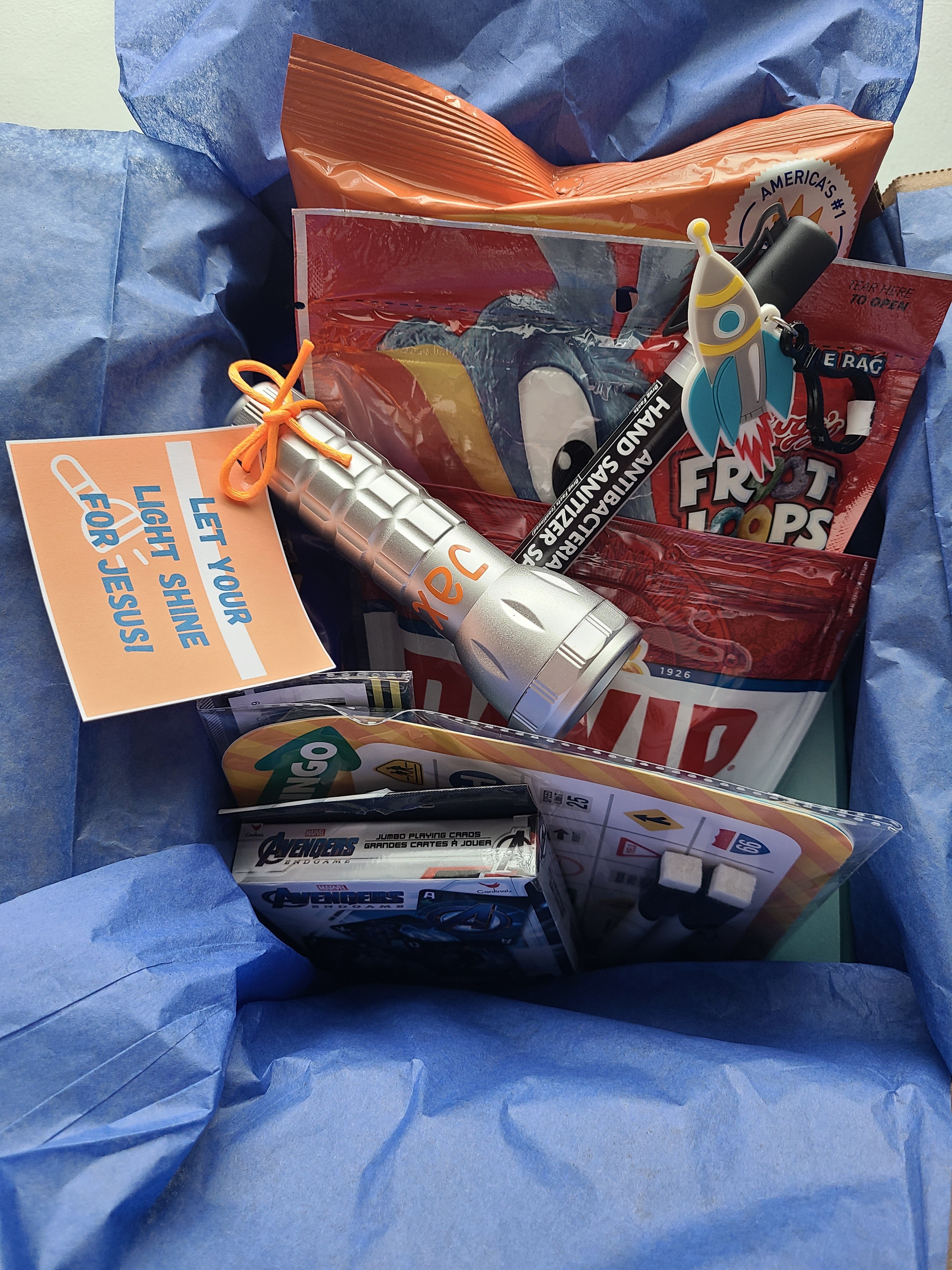 Summer camp care package box lined with blue tissue paper for boys and filled with a flashlight with free printable, hand sanitizer, Froot Loops, Bugles, sunflower seeds, Avenger's playing cards, and road games.