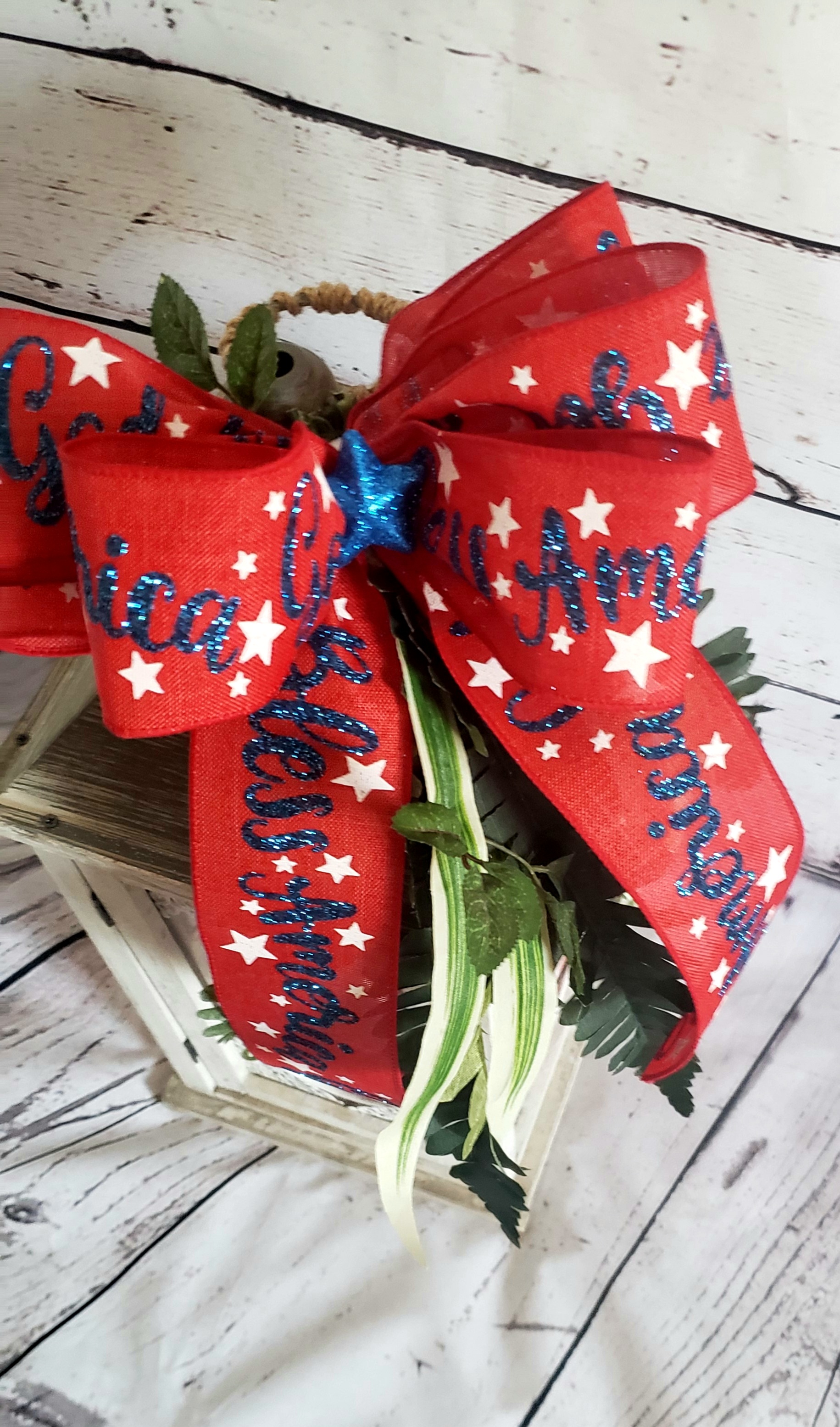 A farmhouse style lantern topped with an easy DIY patriotic decorative swag: greenery draped down the side with a red, white, and blue six-loop bow topping it.