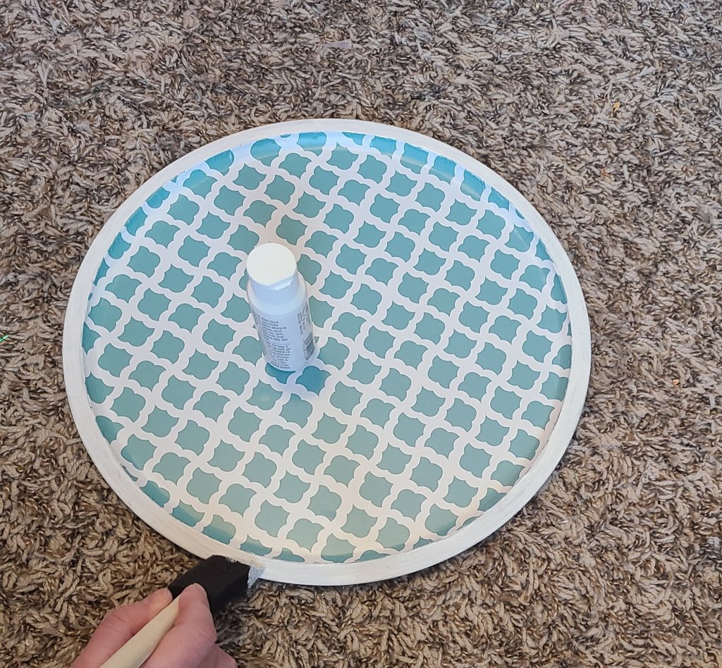 Pizza pan covered with teal and white print contact paper. The edge is being painted white using acrylic paint and a foam brush,