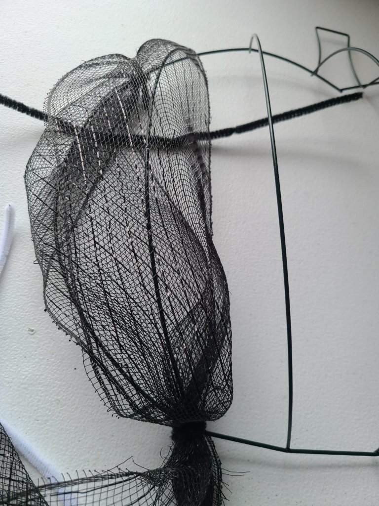 Black deco mesh brought from the bottom of the Halloween pumpkin wreath form up to the top and attached to the left of the first vertical crossbar with a pipe cleaner. This creates a vertical line of deco mesh with some puffiness in the middle.
