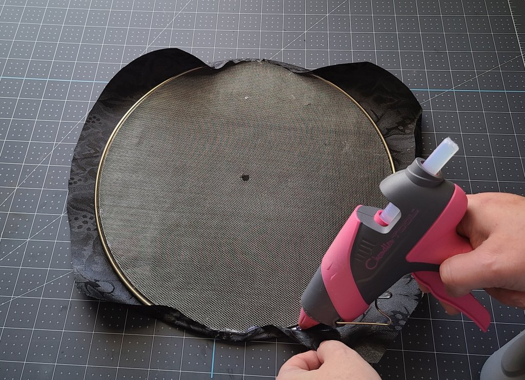 Adding hot glue along the wire of the splatter screen and folding the fabric over the edge. This will be one of the panels on the Halloween splatter screen pumpkin.