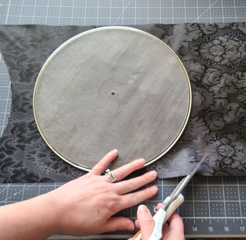 Cutting fabric for a Halloween splatter screen pumpkin. The screen is laid in the middle of a black table runner in the middle height-wise, then leaving about 3" on the sides, scissors are being used to cut around the screen.
