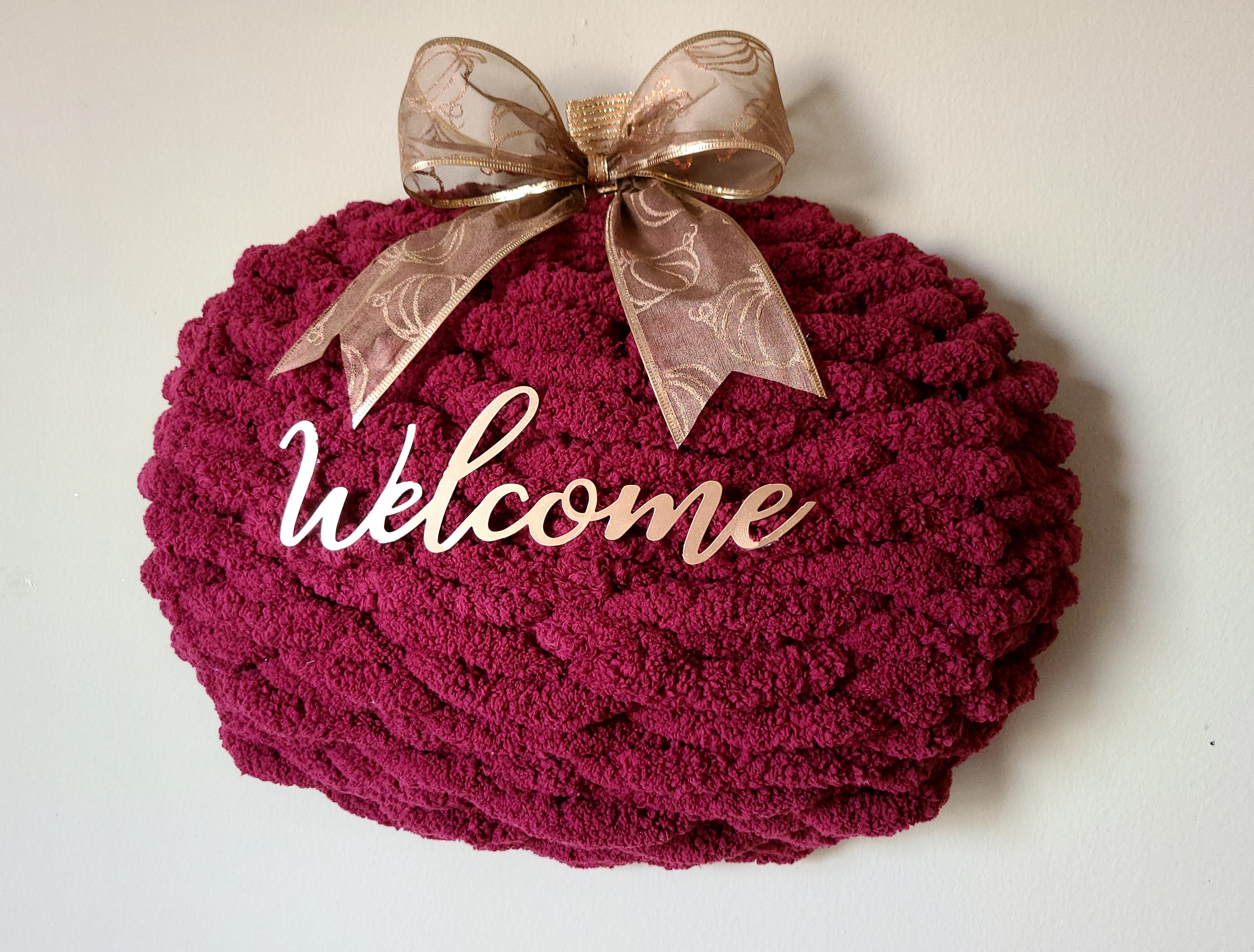 Burgundy chunky yarn pumpkin wreath with copper "welcome" and bow on top hanging on the wall.