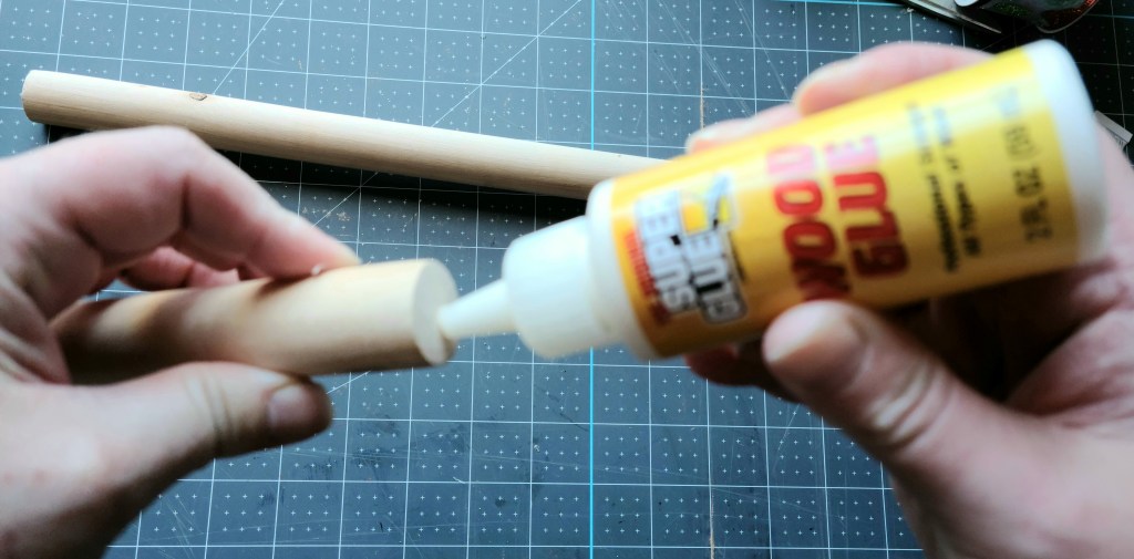 Adding Super Glue brand wood glue to the sanded flat end of the wood plunger handle to attach the two wood pieces to form the pole of the Dollar Tree Christmas mailbox.