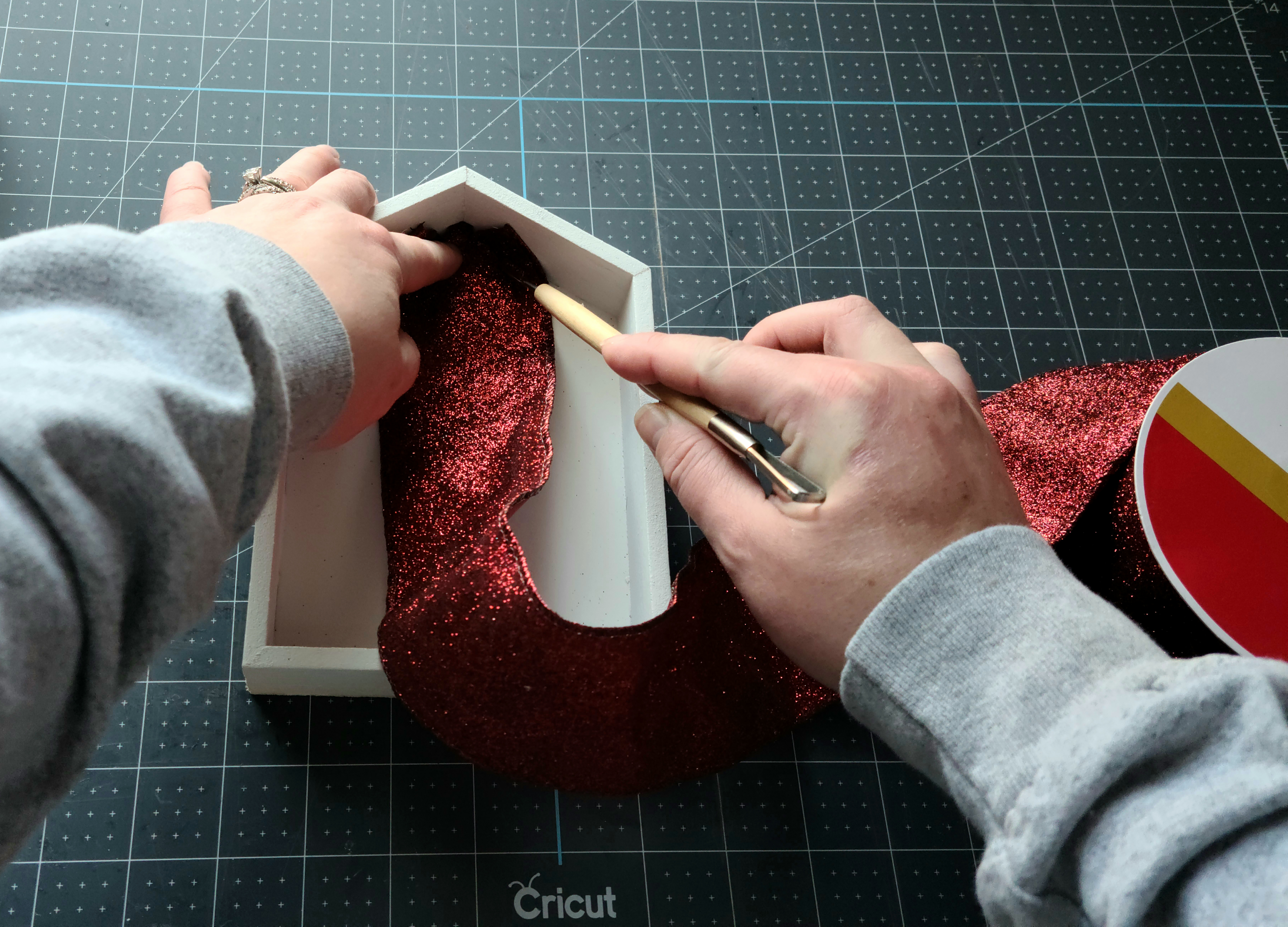 Pressing the end of a 2.5" wired maroon glitter ribbon into the top of the shadow box shaped Dollar Tree Christmas house to create a lines in the ribbon that you can cut along.