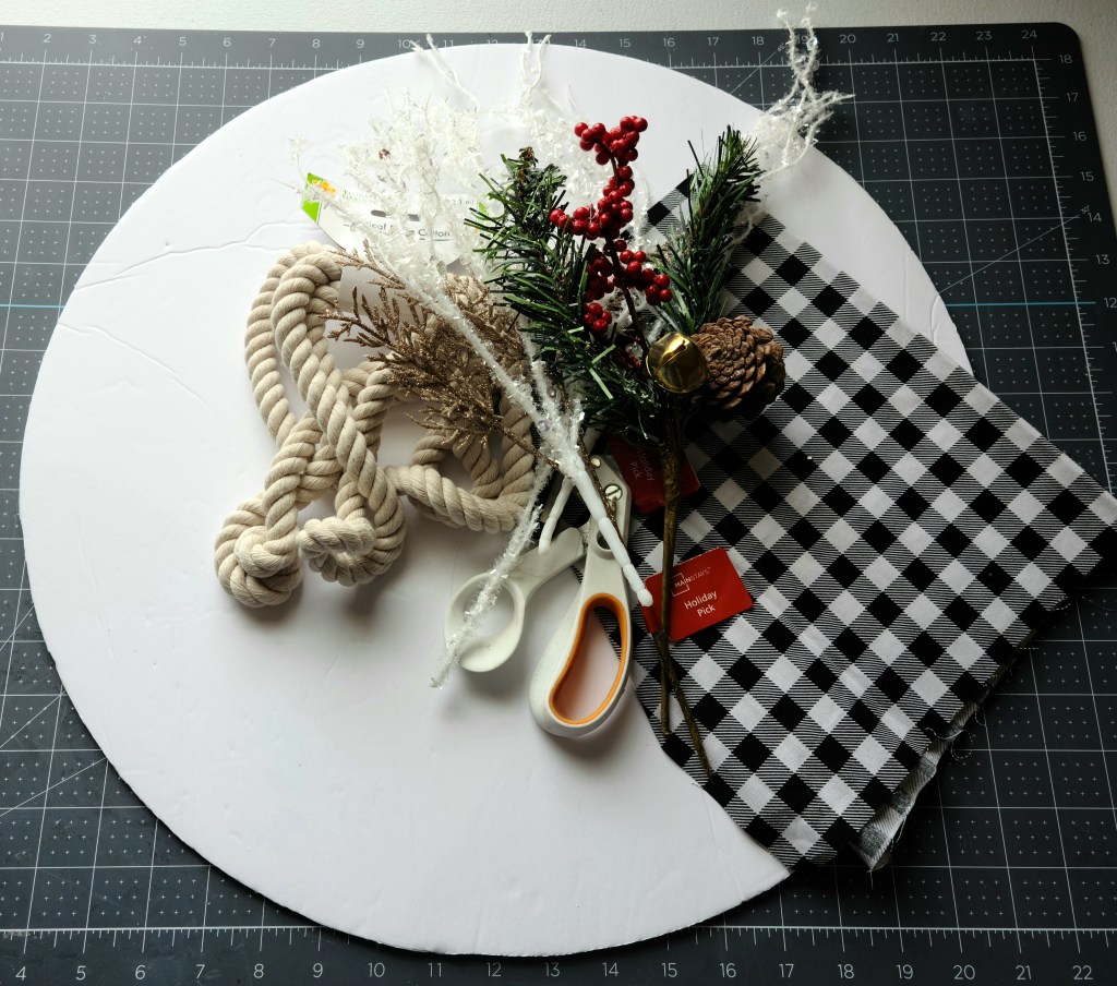 Supplies needed to make a round Christmas sign: foam board cut into an 18" circle, white cotton rope, scissors, floral picks, black and white buffalo check fat quarter of fabric.