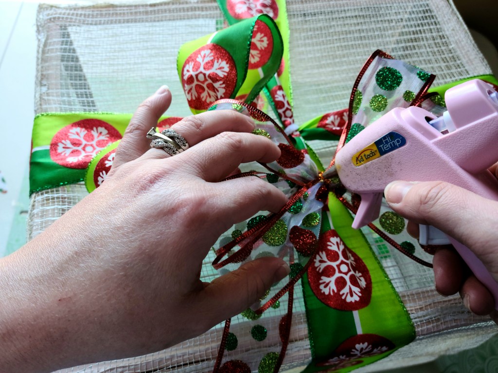 Putting hot glue on the middle of a six loop bow made out of 1.5" sheer ribbon with red edges and red, green, and light green glittered circles.