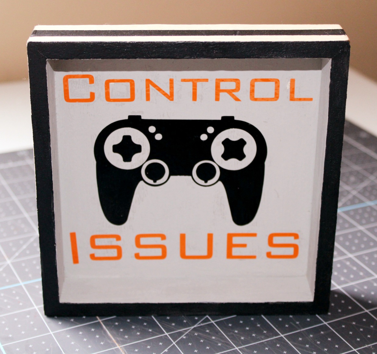 DIY gamer room sign completed. It's a 5x5 sign that is similar to a shadowbox. The outer frame is light colored wood with a black stripe painted in the middle, the front is black, the insides are painted gray, and there is a picture of a gaming controller in black with the word "control" issues in orange above the controller and "issues" in orange underneath.