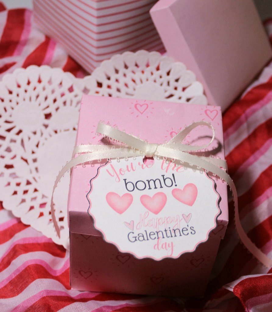 Valentines Day gift for friends: pink box with hearts with a cream ribbon wrapped around it which is attaching the "You're the bomb!" gift tag on.