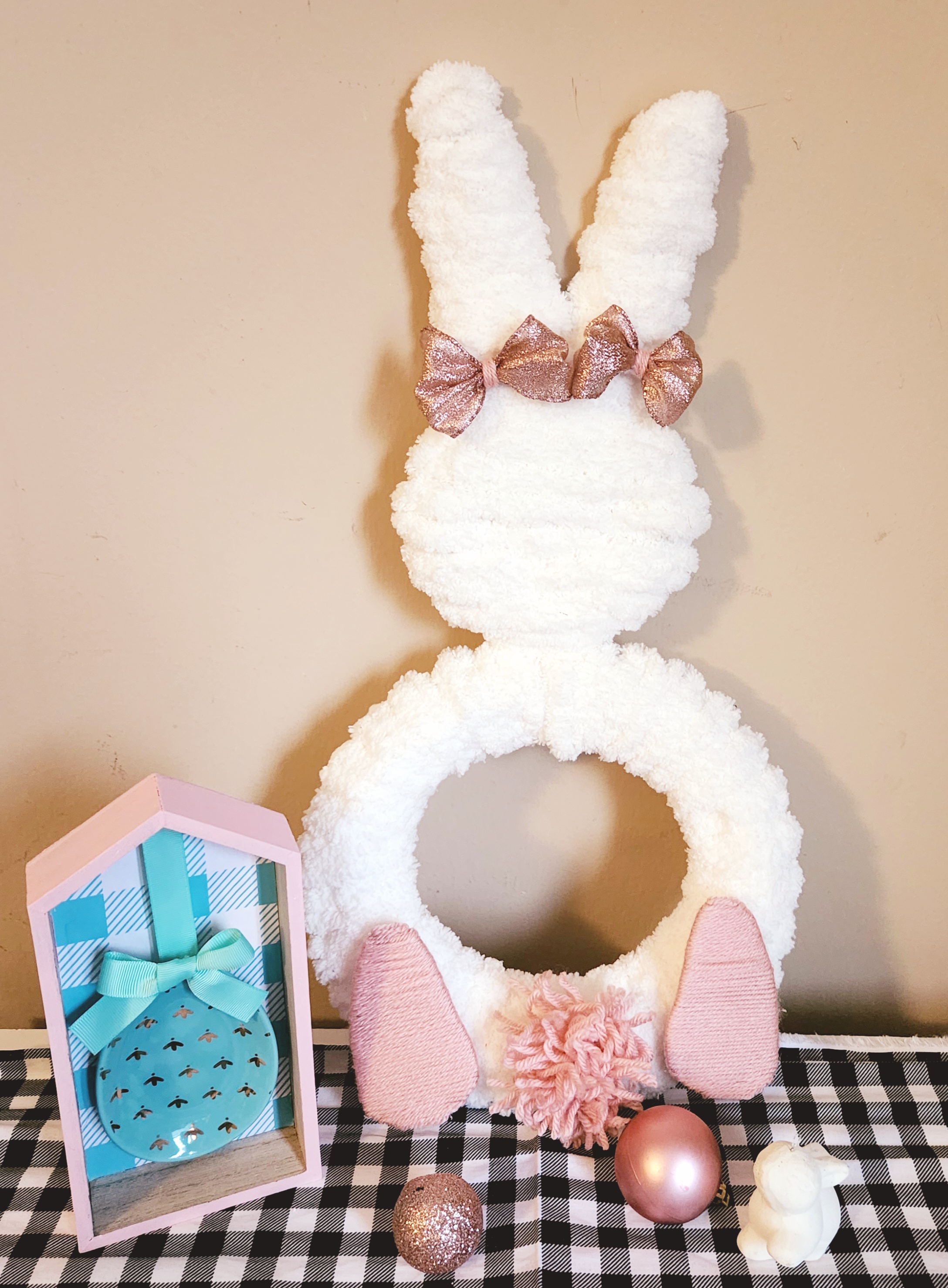 bunny wreath: Completed bunny wreath wrapped with chunky white yarn covering the head completely with the body wrapped like a regular wreath form having an open middle, there are two feet facing outward on the bottom, a pink pompom tail, and two metallic pink bows on the ears.