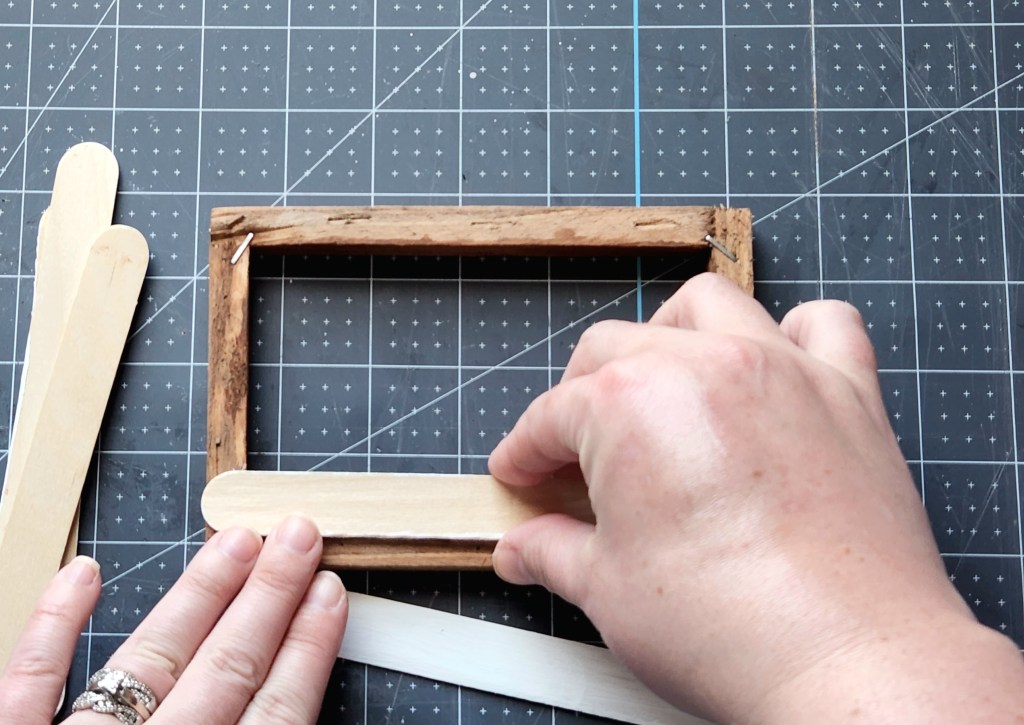 Placing a painted popsicle stick on top of the hot glue on the back of the Dollar Tree canvas wood frame.