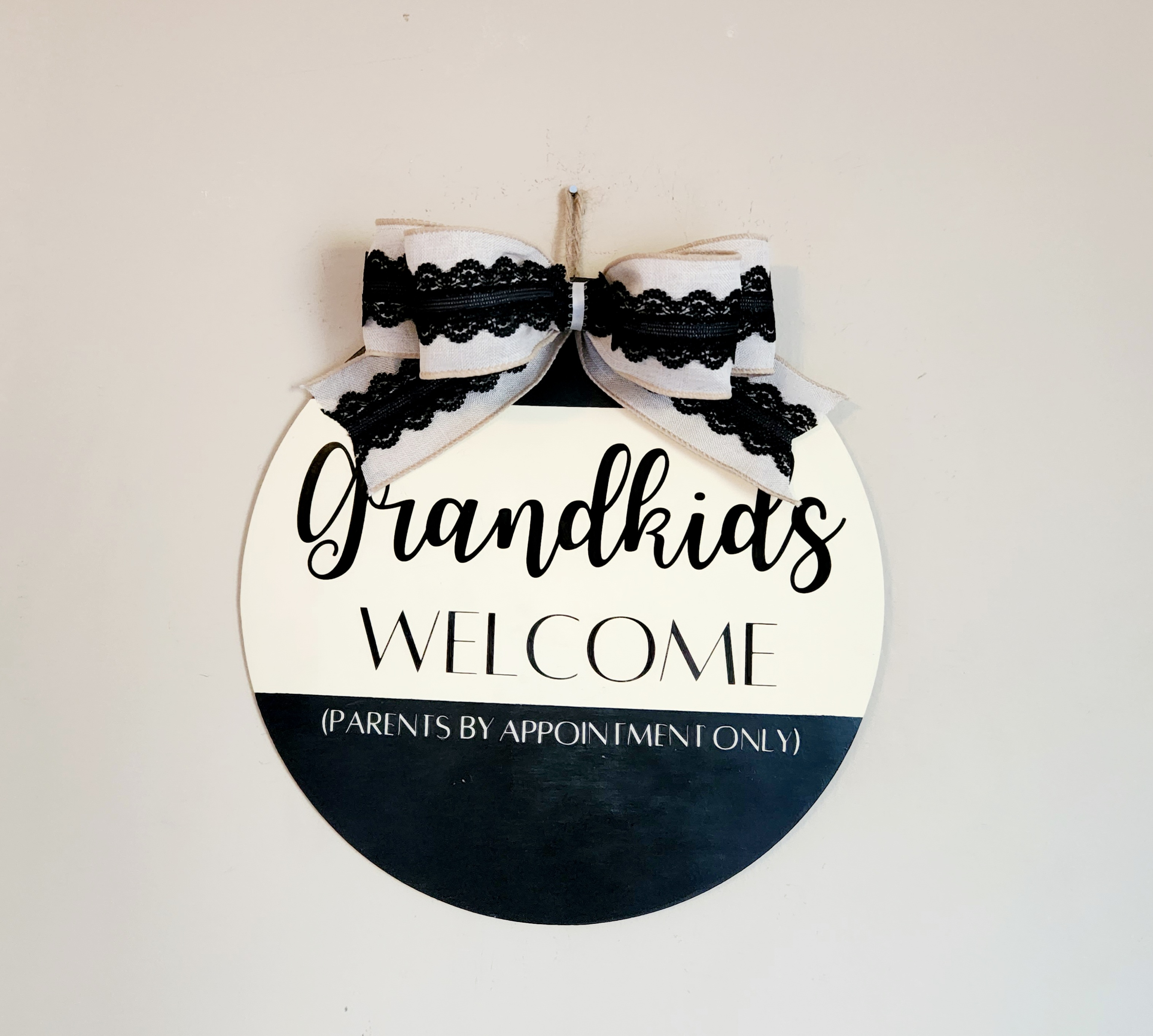 Round sign with color blocks of black, ivory, and black at the bottom saying in the middle "Grandkids Welcome (parents by appointment only)" topped with a khaki linen and black lace bow.