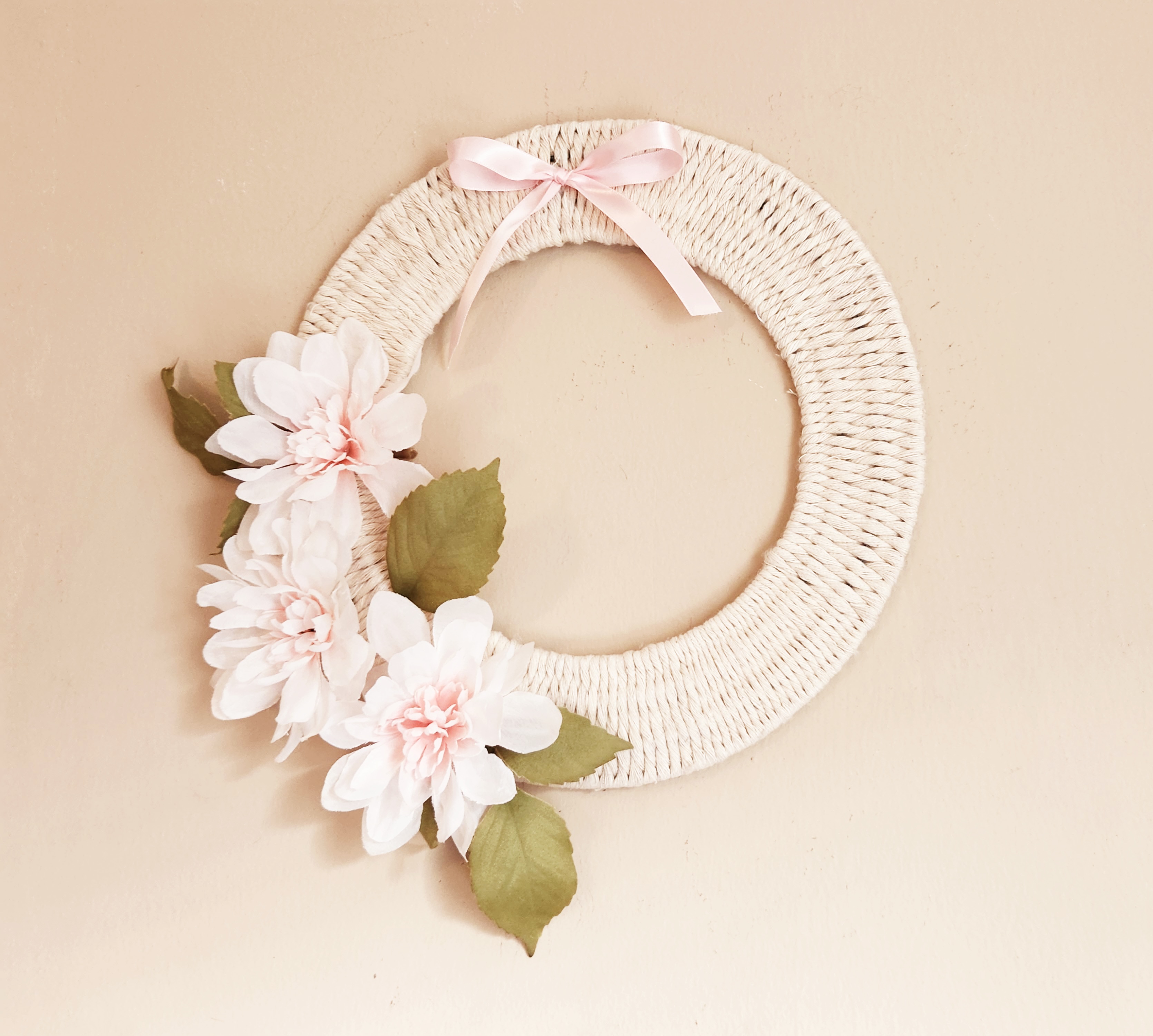 Woven rope wreath made with white cotton rope woven onto a wire wreath form with pink dahlias across the lower left side and a pink satin ribbon on the top.