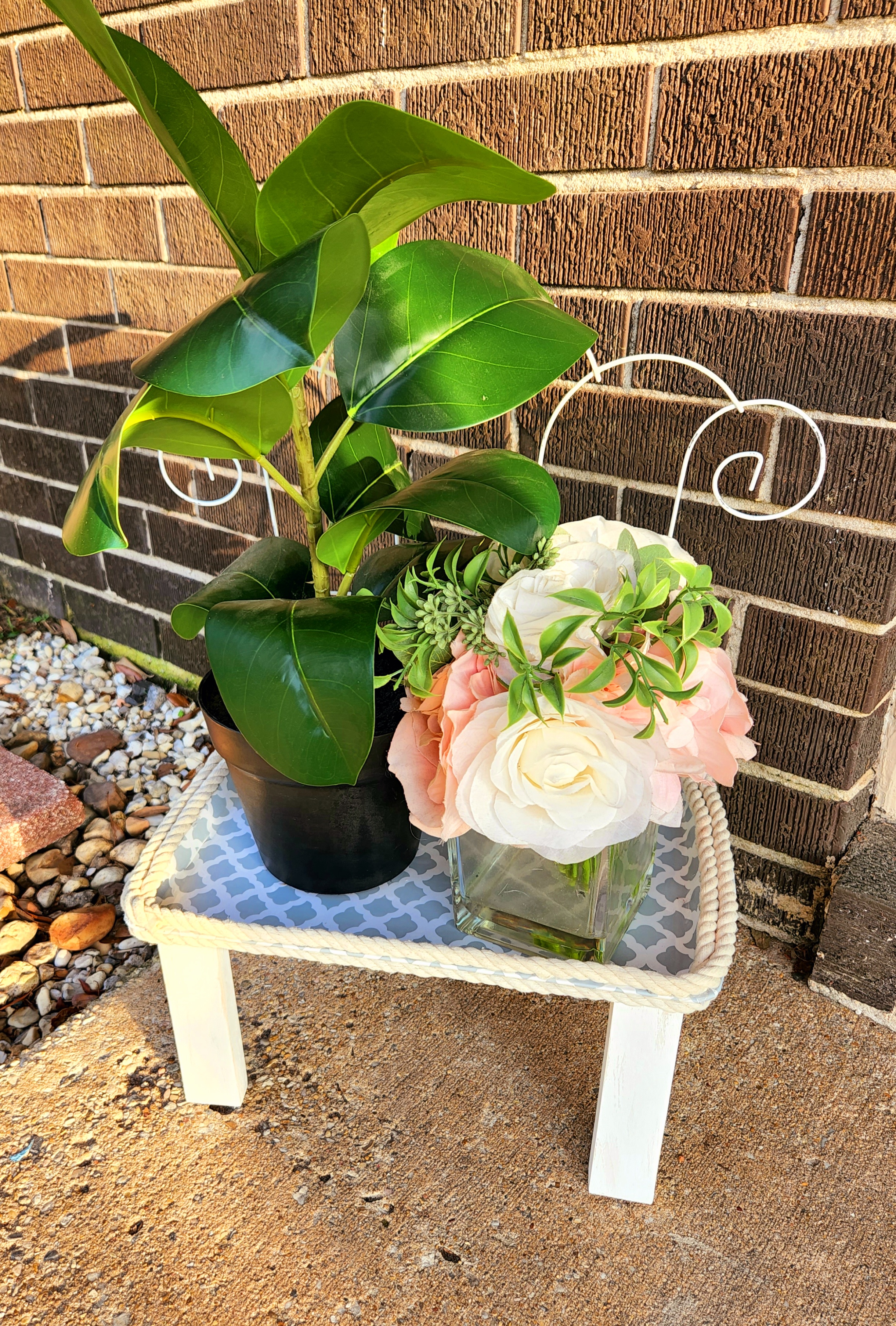 Outdoor plant stand on my porch with a plant and vase of flowers placed on it.