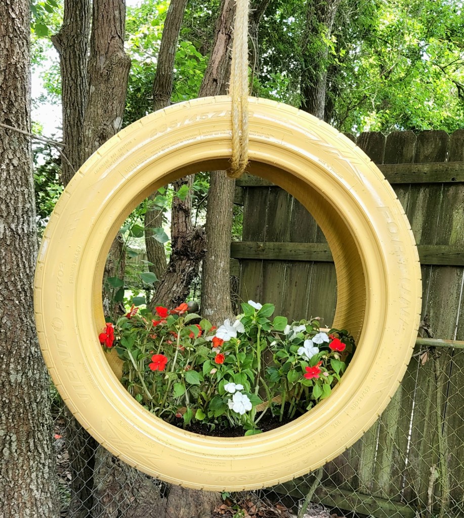 Completed tire planter. Tire painted yellow with red and white impatients planted inside and hanging from a tree with sisal rope.