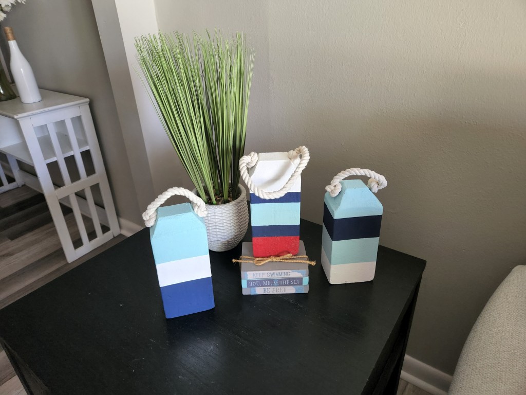 3 nautical buoys with red, white, blue, and robin's egg stripes painted it on them and a vase with sea grass in them next to them on a tabletop.