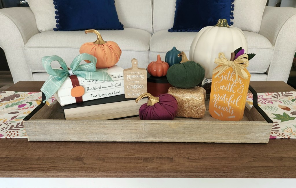 Early fall vignette: Tray on a fall table runner with stacked books, pumpkins, a small hay bail, white pumpkin, small decorative cutting board, a small orange porcelain pumpkin next to a small teal one.