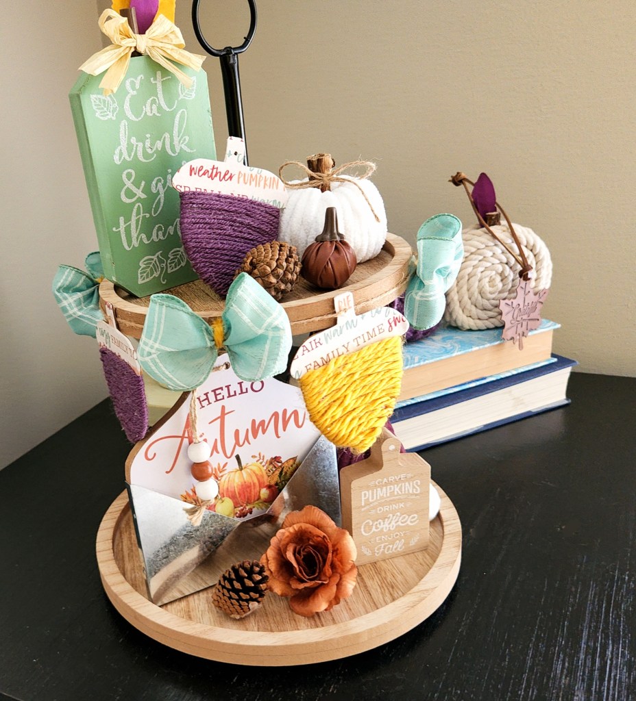 Acorn garland added around the top tier of a two-tier tray filled with fall items, yarn pumpkins, small pinecones, small fabric and leather pumpkins, a "Hello Autumn" pocket sign, and a wood mint pumpkin.