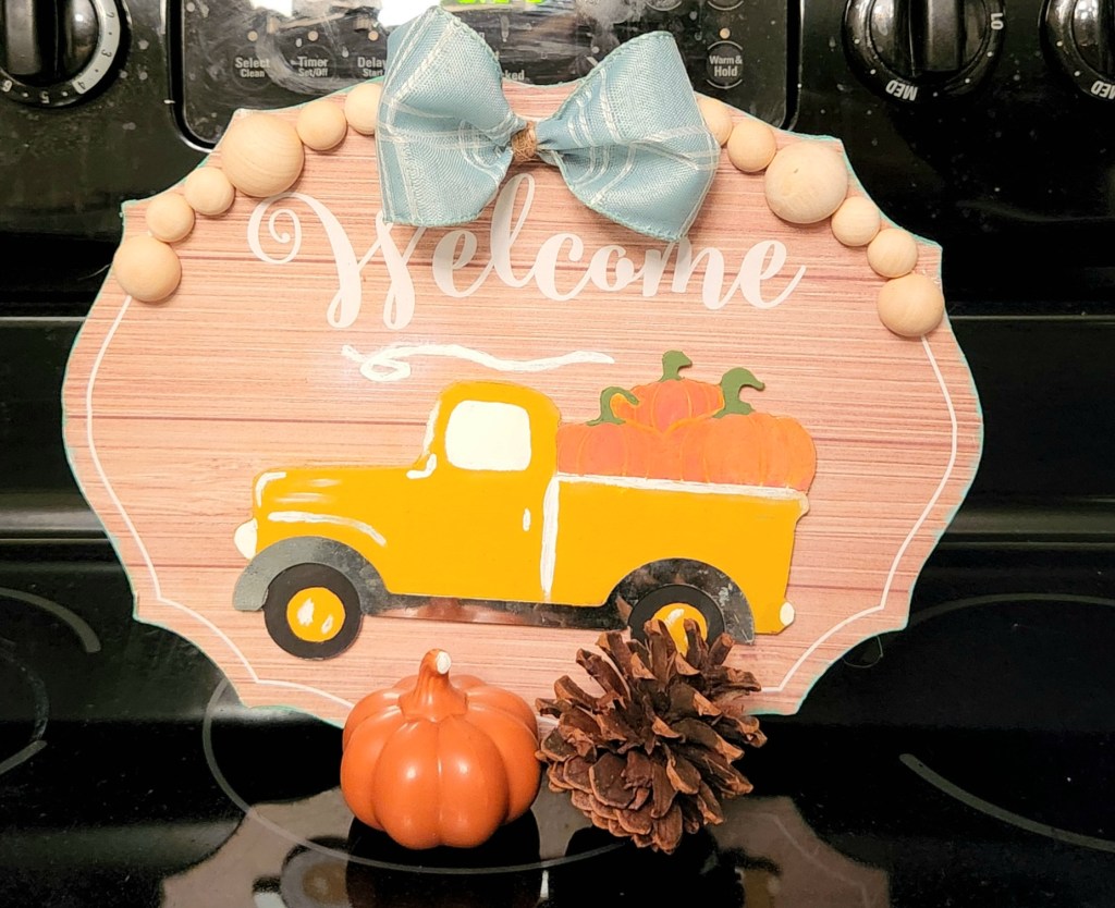 Fall Dollar Tree sign upcycle- says, "Welcome" in the middle with a yellow farmhouse truck carrying pumpkins and a mint bow and wood beads across the top.