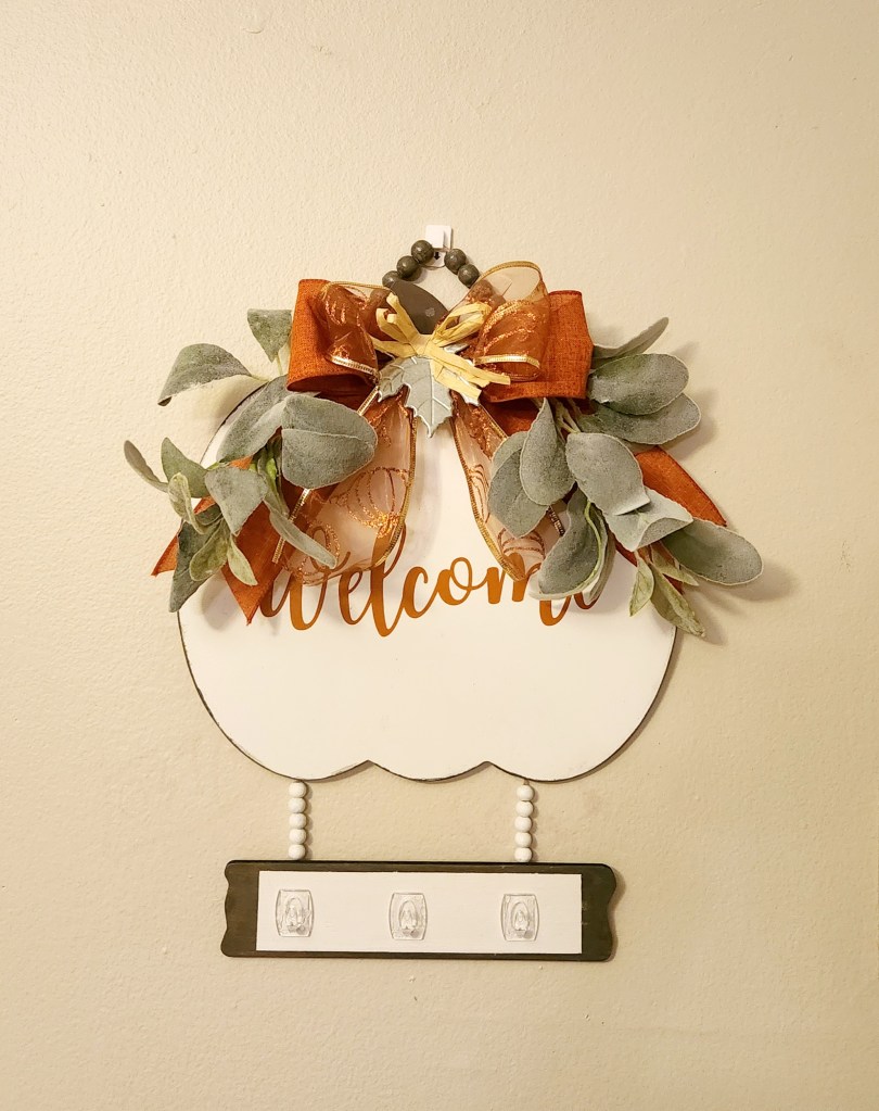Pumpkin shaped keyholder with "welcome" written in orange spice and lamb's ear branches on each side and an orange bow with a metal leaf in the middle of the bow.