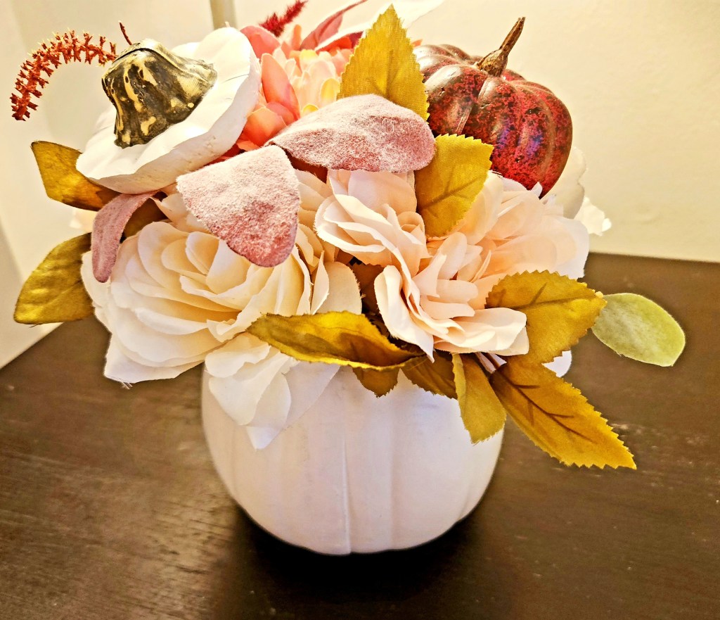 White pumpkin turned into a vase filled with fall florals in pink and white with a burgundy pumpkin pick inside.