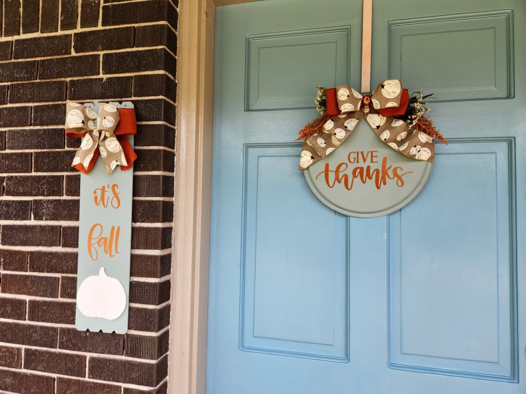 Fall pizza pan wreath hanging on a door, saying, "Give thanks," with two bows at the top, one orange and one burlap with white pumpkins, and orange and white baby's breath greenery at the top. A matching sign saying, "it's fall," in orange with bows on top and a white pumpkin at the bottom, hanging next to the door.