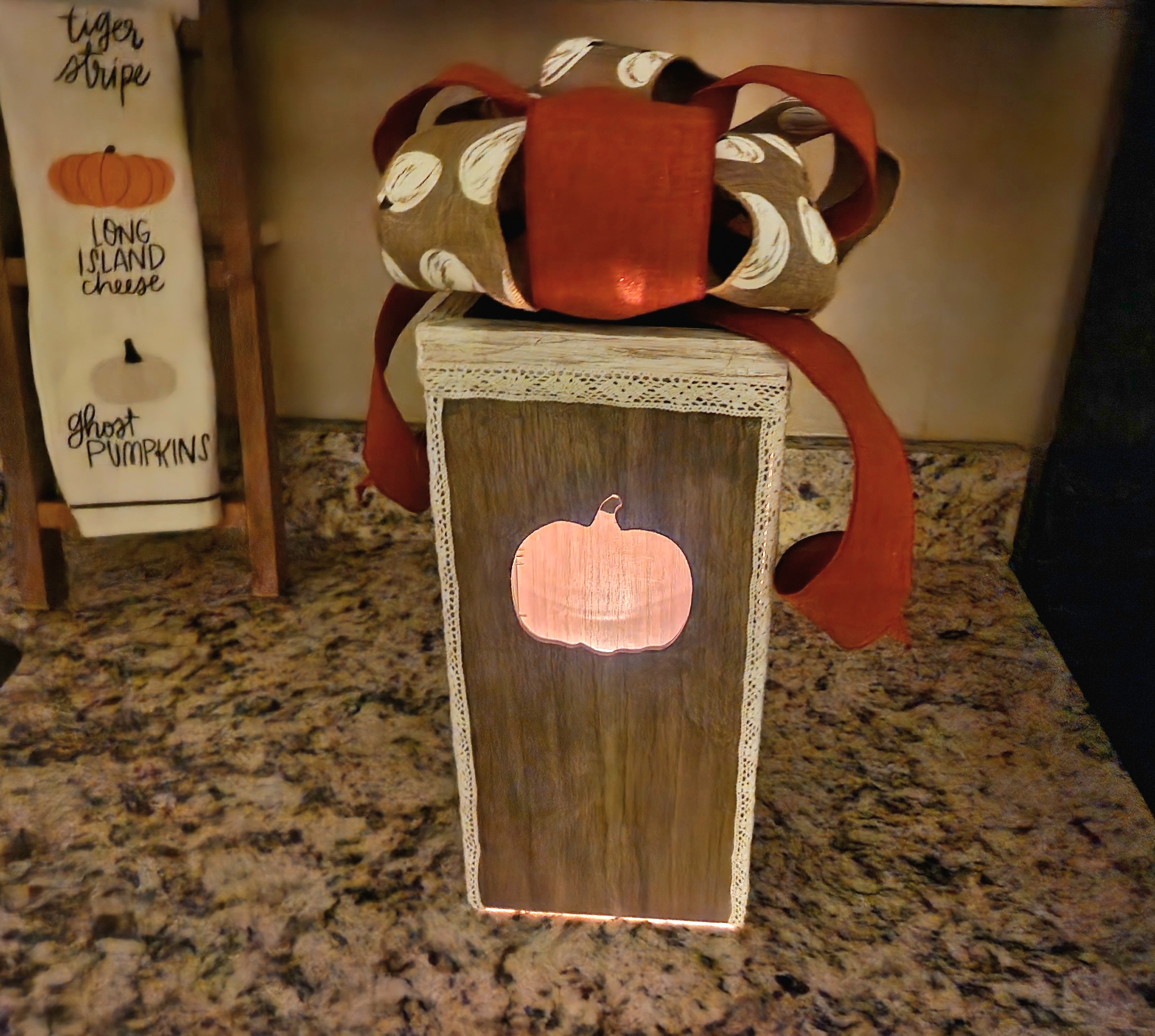 DIY fall lantern with pumpkin cutouts on the sides and an orange ribbon and white pumpkin print bow on top. Lights are off and a candle is lit inside.