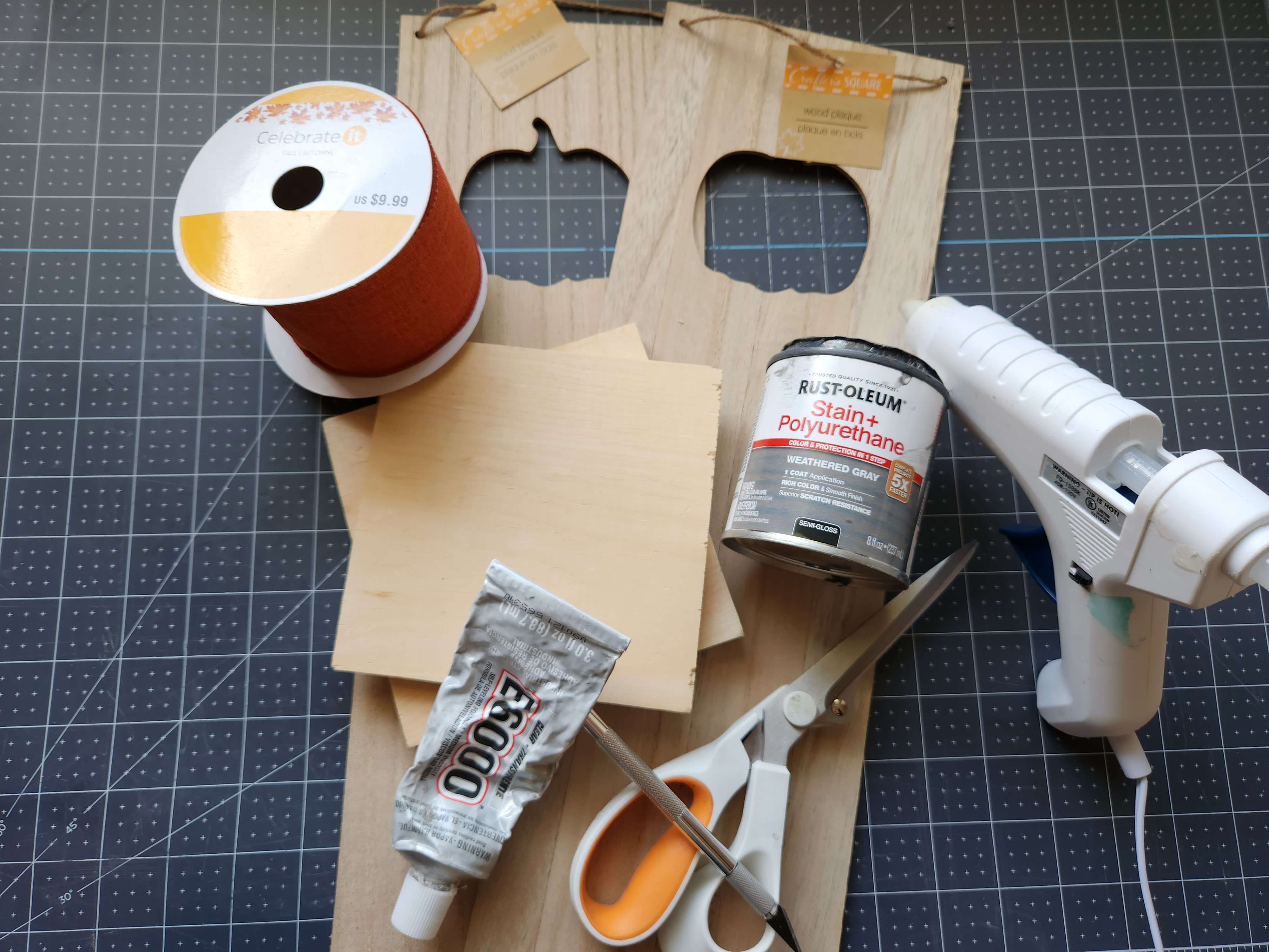 Supplies for a fall lantern: 3 wood panels with pumpkin cutouts, weathered gray wood stain, E6000, hot glue gun, scissors, exacto knife, and orange ribbon.