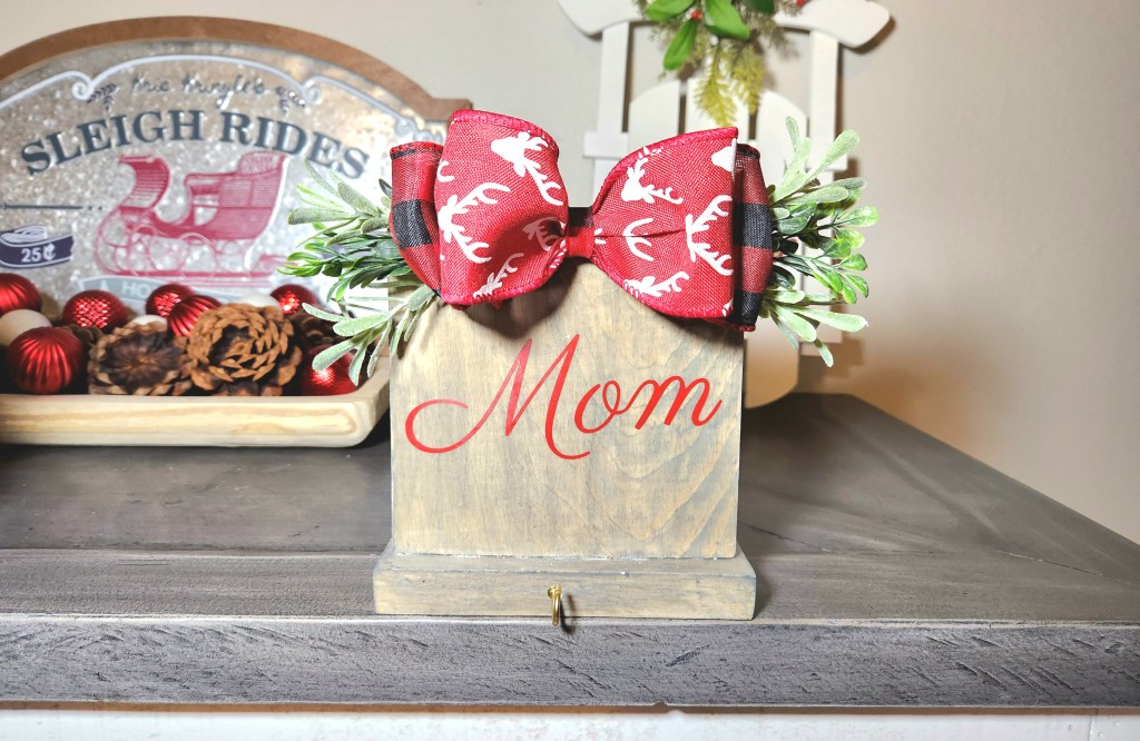 Christmas stocking holder with "Mom" on weathered gray stained wood, gold hooks at the bottom, and topped with greenery and Christmas bows.