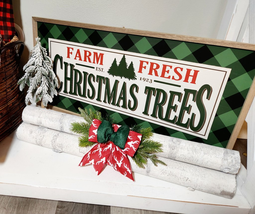 Three faux birch logs bound together with a red reindeer & green linen bow & faux greenery behind the bows. Behind the faux birch logs is a down-swept fir flocked faux Christmas tree and a large green & black buffalo check sign that says, "Farm fresh Christmas trees Est. 1923."
