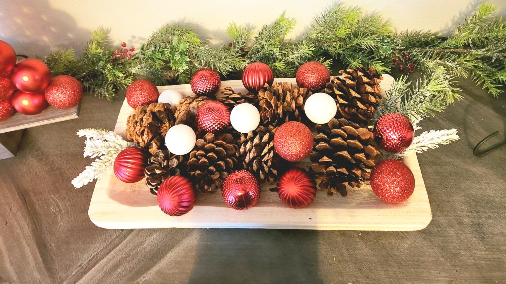 A dough bowl filled with pinecones, red ball ornaments, tiny white ball ornaments, and a few pieces of greenery on each end.