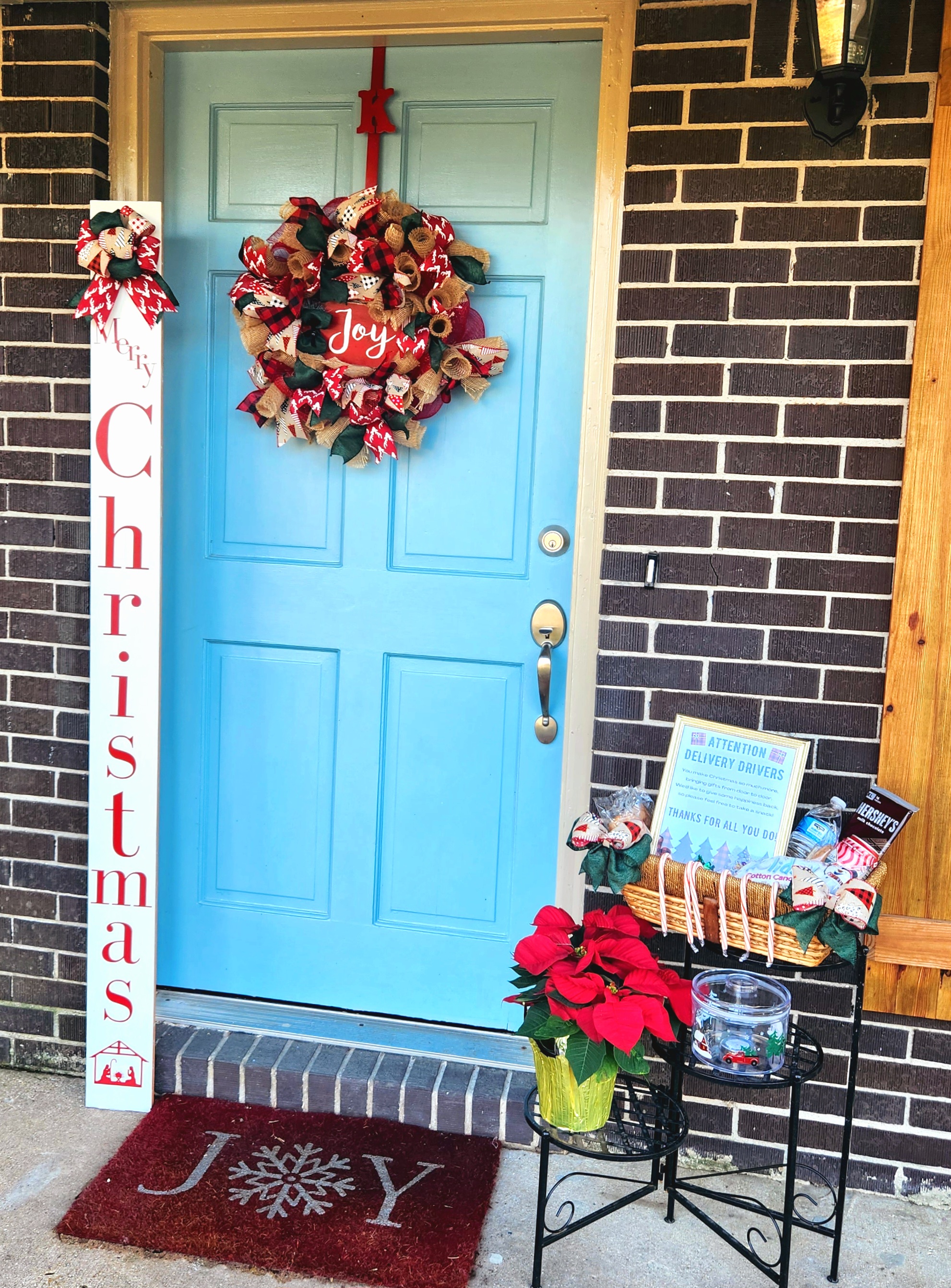 Our Christmas decorated porch with a "Joy" wreath on the door, a "Merry Christmas" porch leaner next to it, and a Christmas delivery driver printable sitting inside a basket filled with drinks, candy, and other snacks. The basket is on a plant shelf with a poinsettia on the lower shelf.