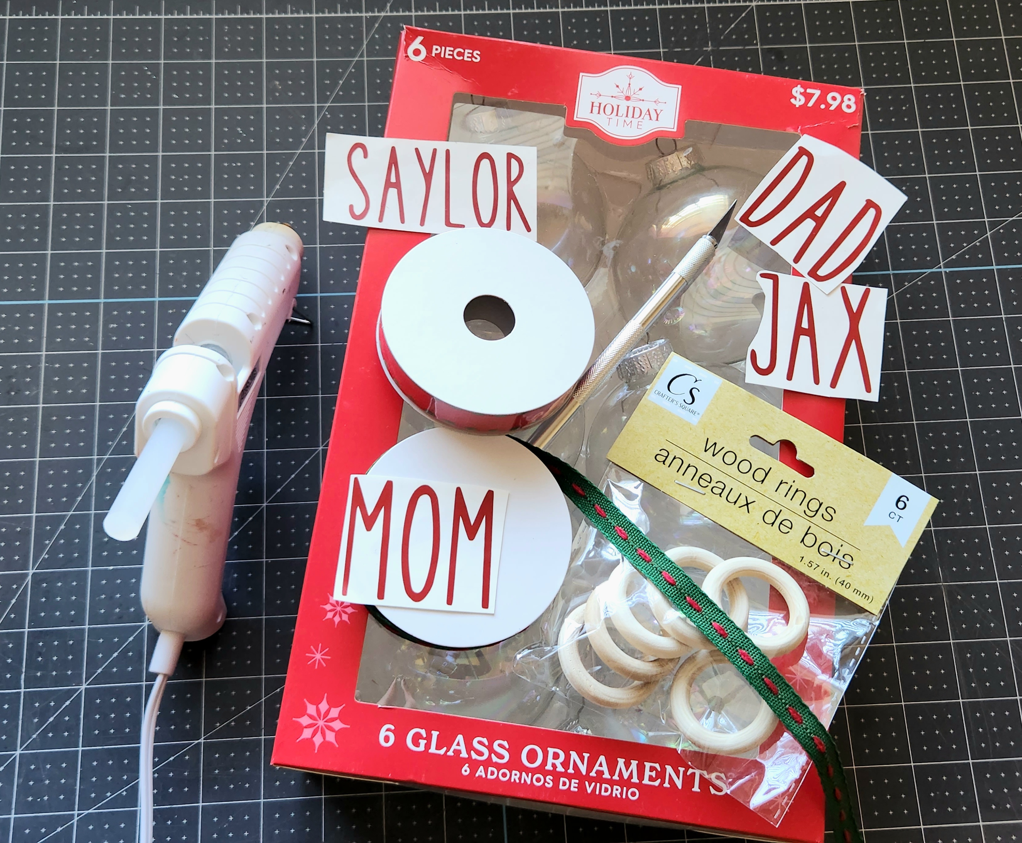 DIY ornament glass supplies: clear glass ornaments 6 pack, vinyl names, red and green ribbon, wood rings, exacto knife, and a hot glue gun with glue.