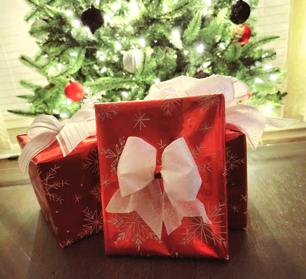 Christmas presents wrapped with red wrapping paper with white snowflakes with different embellishments on each one. The gifts have white bows made with different textures of ribbon on top. They are sitting in front of a white lighted Christmas tree.