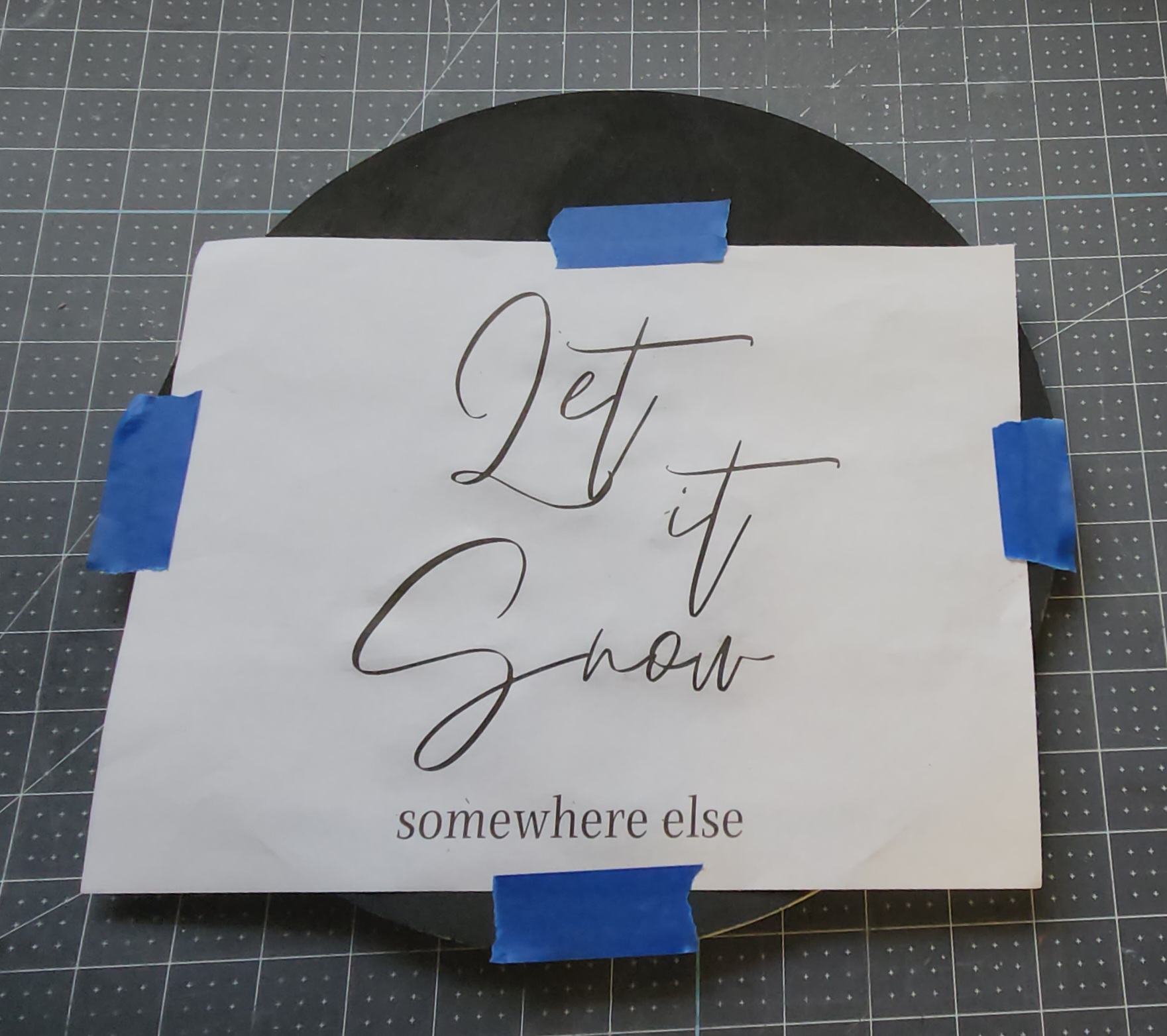 "Let it snow somewhere else" printed on paper and taped to a round piece of black wood with painter's tape.