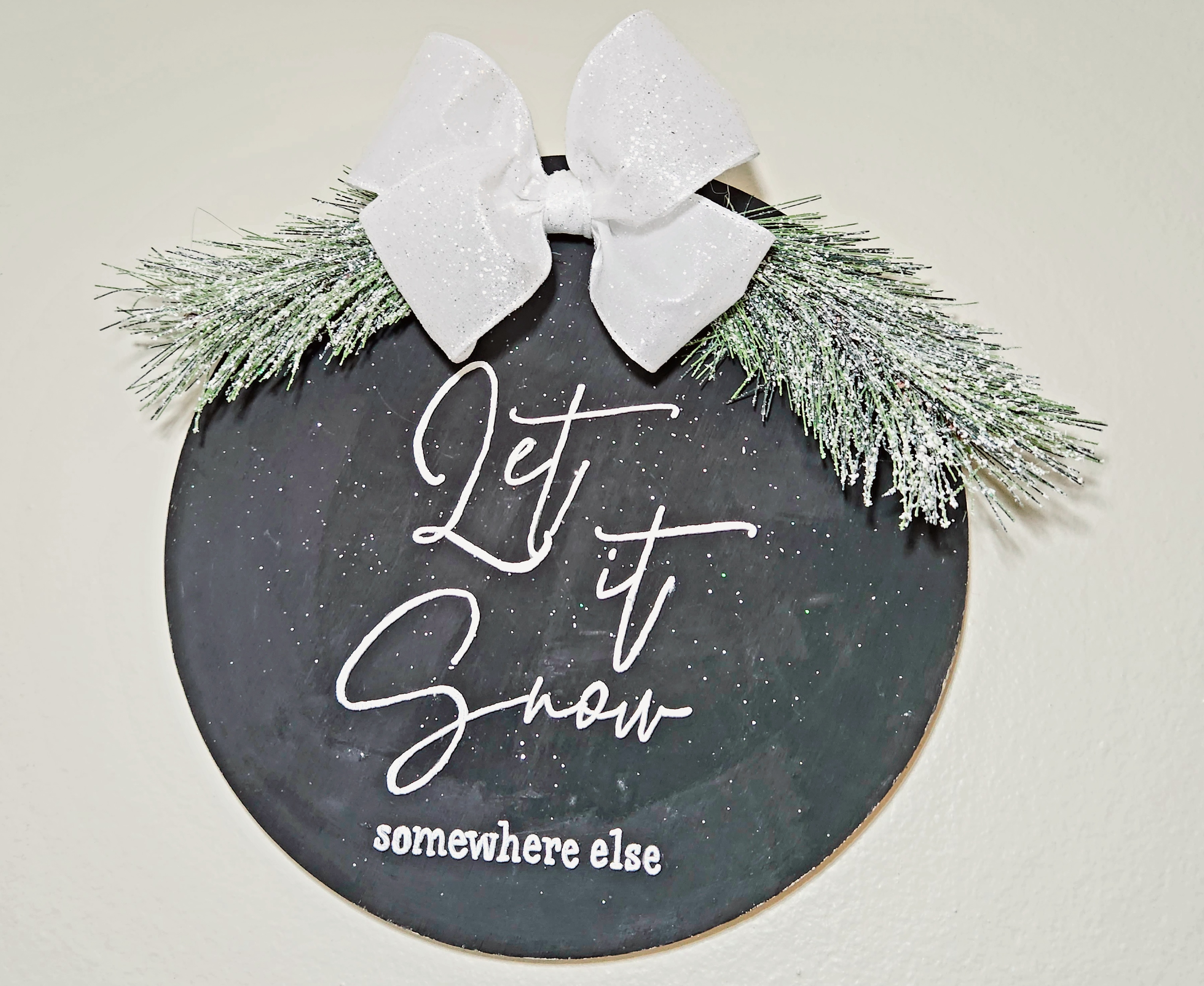 Black winter door hanger that says, "Let it Snow, somewhere else," with a white glitter bow and flocked greenery at the top.