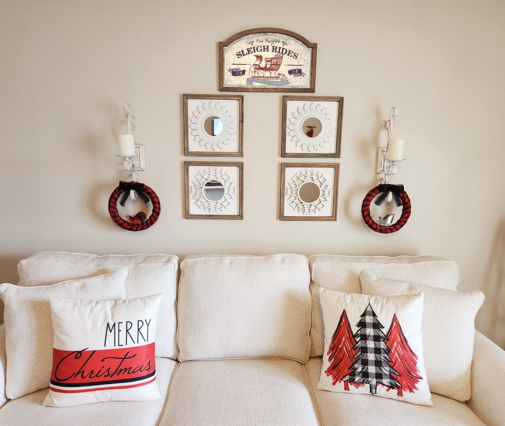 Buffalo check Christmas wreaths hanging from sconces with four decorative mirrors in between the sconce and a galvanized metal sign above them. They are hanging above a cream couch with decorative Christmas pillows, one with black buffalo check and red trees on each side of it, and the other with "Merry Christmas" written on it with Rae Dunn and script fonts with red and black stripes toward the bottom.