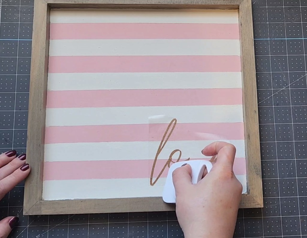 Transferring the word "love" in gold glitter vinyl on the bottom left quadrant of the Valentine's Day sign by tracing over it with a scraper.
