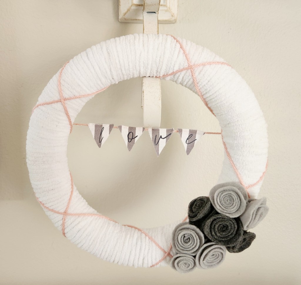 Valentine's wreath covered with white chenille yarn with dusty mauve yarn wrapped on top in an X pattern, light and dark gray felt rosettes in the bottom right corner, and a gray and white banner across the middle with "love" on it.