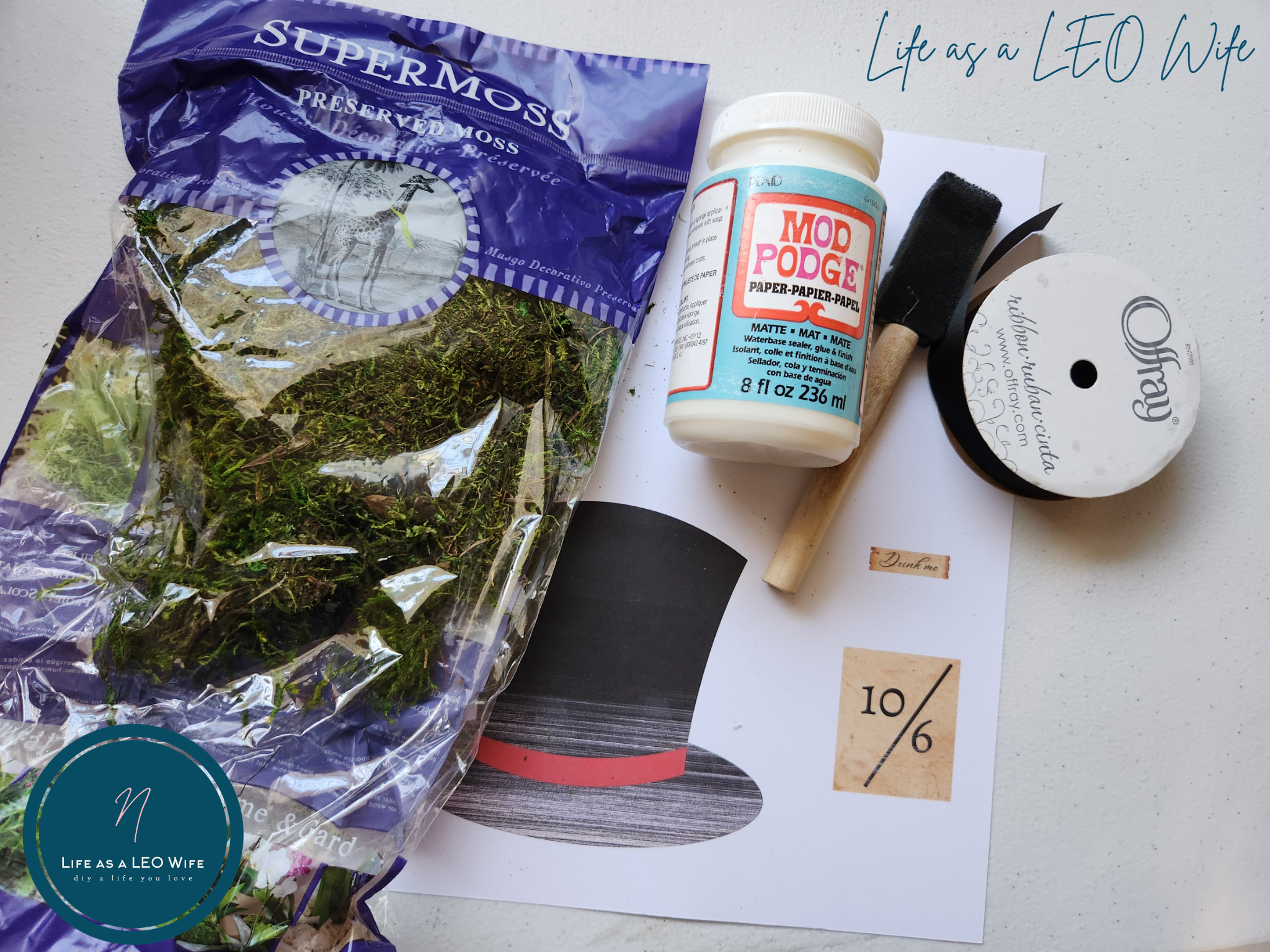 Supplies for a DIY Mad Hatter hat to place on the Alice in Wonderland tea tray: top hat printed on cardstock, 1/2" black ribbon, foam brush, Mod Podge, and preserved moss.
