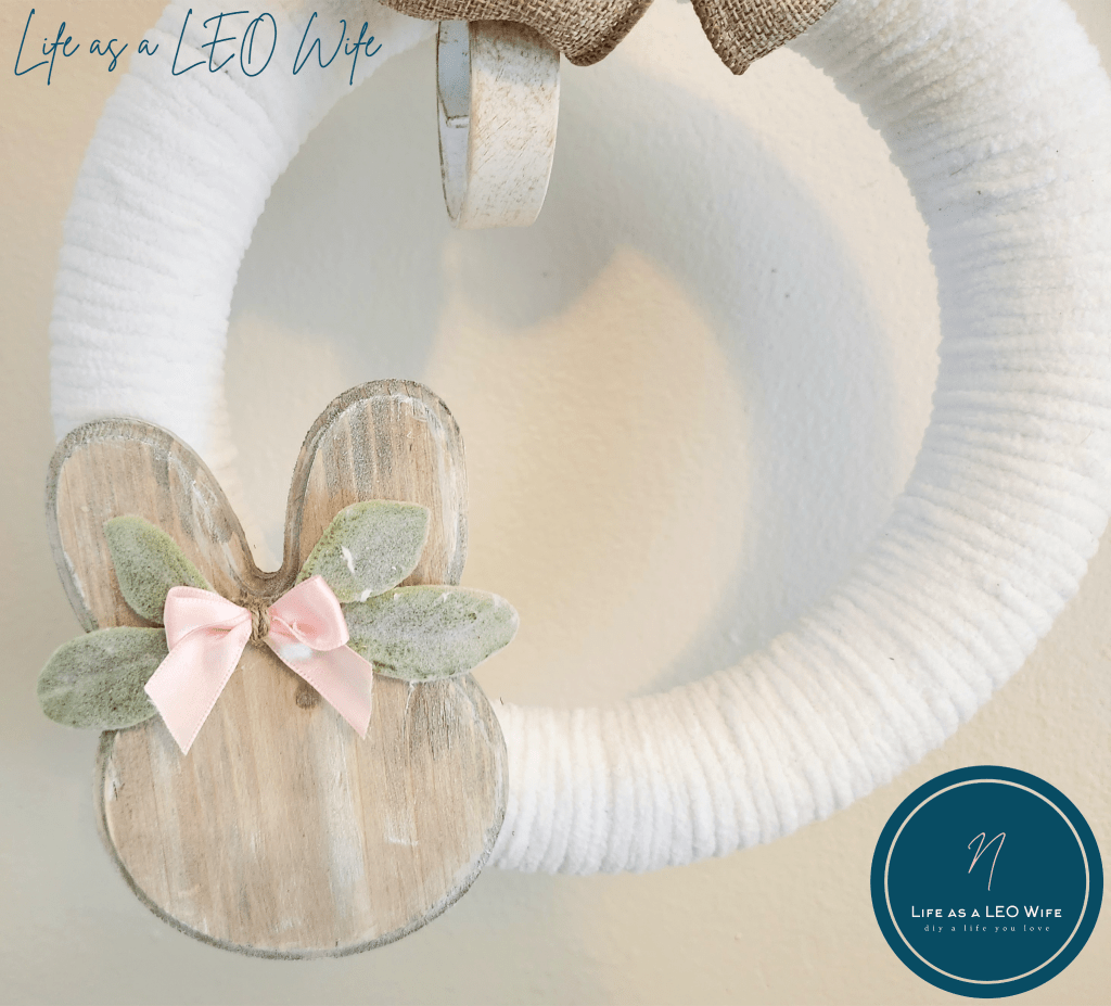 Spring wreath wrapped in white chunky yarn with a tan linen bow and a wood bunny stained with weathered gray stain and a pink bow with lamb's ear leaves behind it.