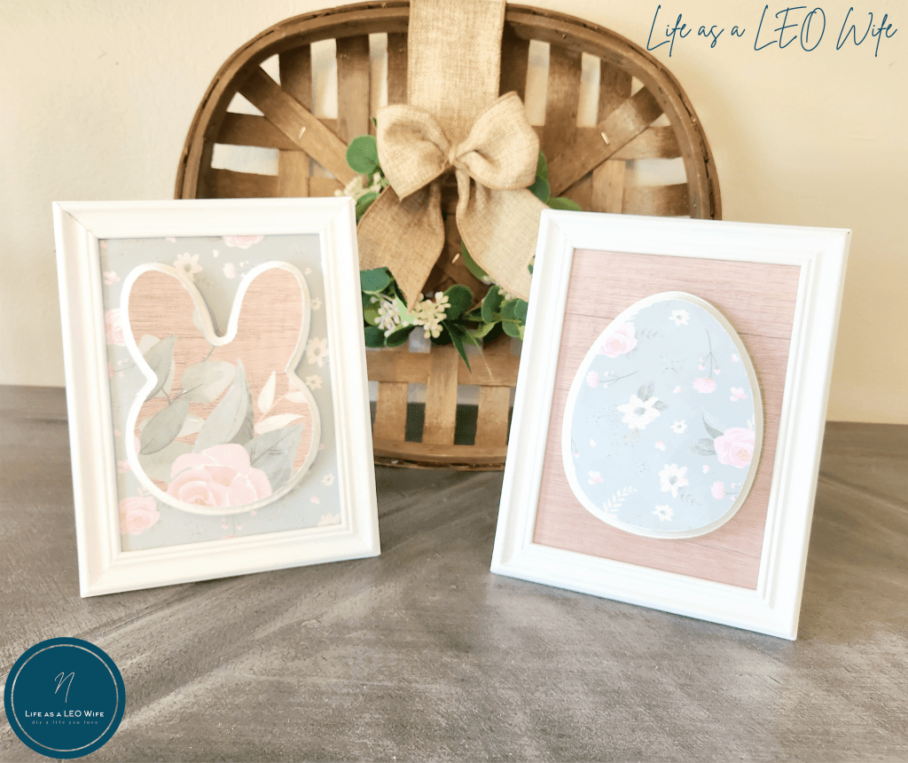 White 5x7 fame with mint, white, and pink cardstock inside the frame with a white wood bunny covered with wood print cardstock paper with mint and pink cardstock paper attached to the front with Mod Podge. Next to it is a framed wood Easter egg with cardstock paper opposite to the bunny frame: wood pattern in the frame and floral paper on the egg. Both the framed bunny and Easter egg are in front of a tobacco basket with a wreath hung inside it.