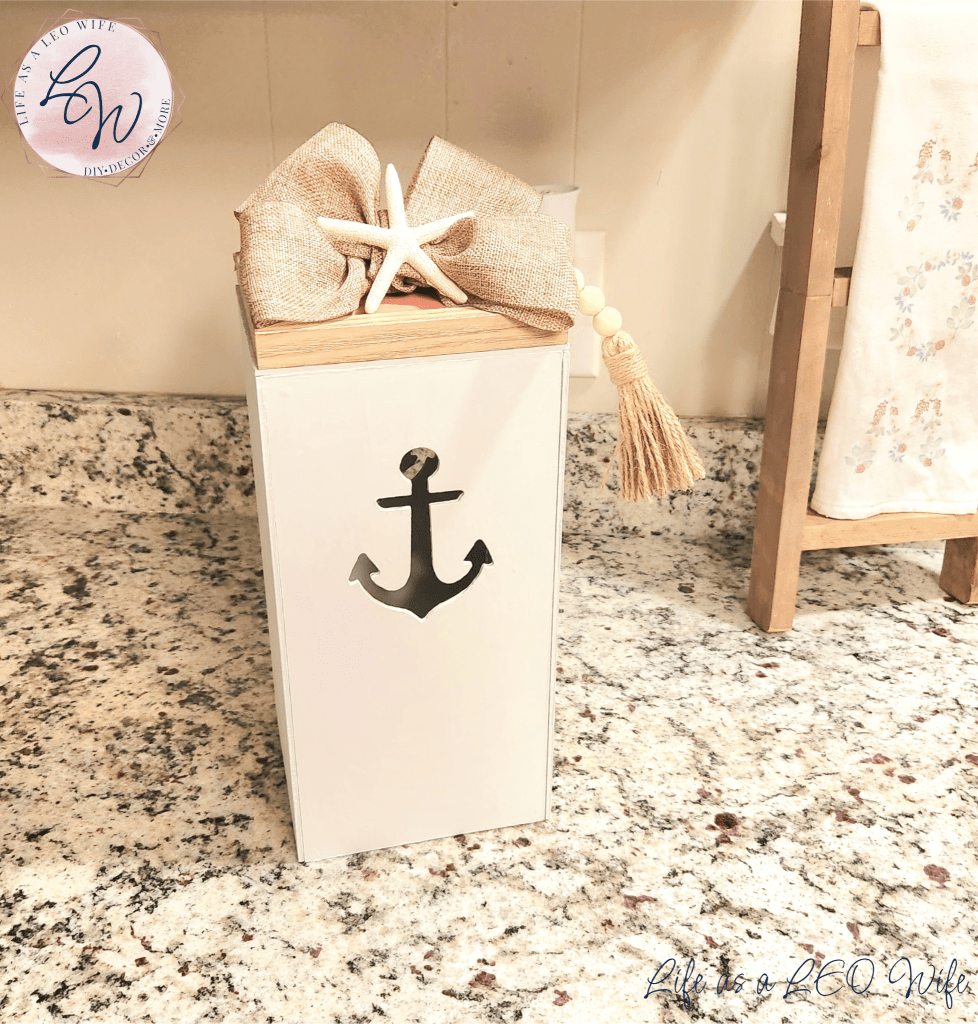 DIY coastal farmhouse lantern painted white with an anchor cutout on two sides, topped with a neutral colored frame with a coral interior and a khaki linen bow with a starfish in the center.