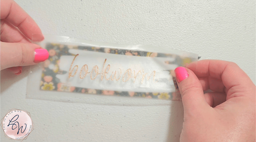 Placing gold glitter vinyl "bookworm" onto white brush stroke in white vinyl on top of floral print DIY bookmark made from Dollar Tree cutting mat.