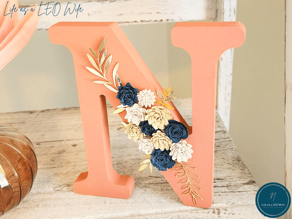 Wood letter N painted coral with different styles of paper flowers in the center in navy, light peach, and tan, with metallic gold leaves extending from behind them.