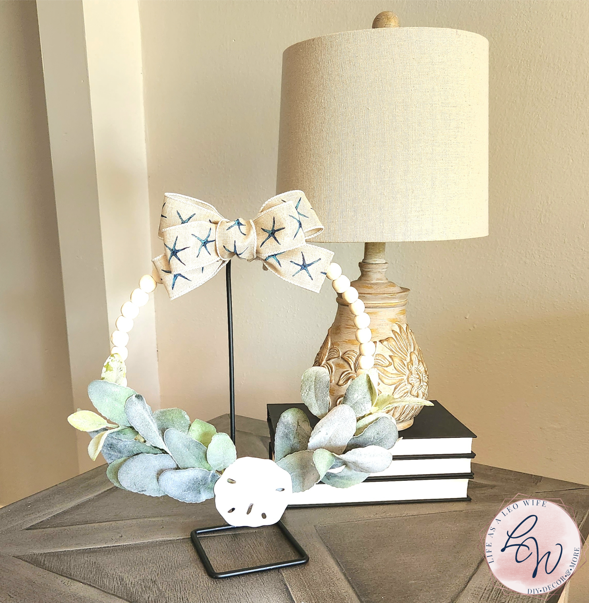 Coastal wood bead wreath with lamb's ear greenery across the bottom, a sand dollar in the bottom center, and topped with a starfish print bow, hung on a wreath stand.